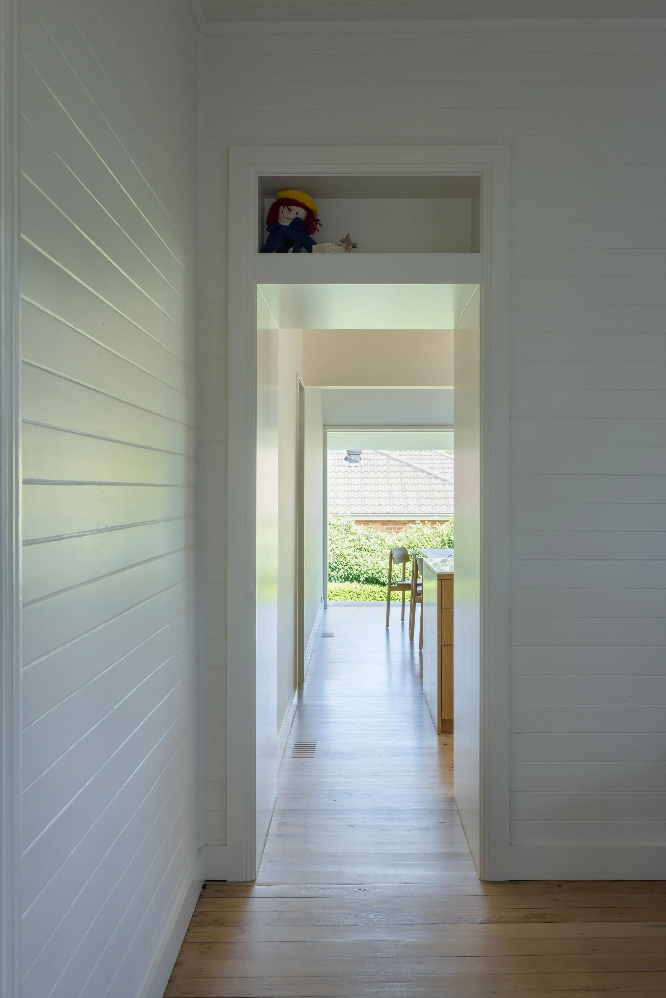 A hallway with white wooden walls leading to a sunlit room with a view of the outdoors, a wooden table, and chairs.