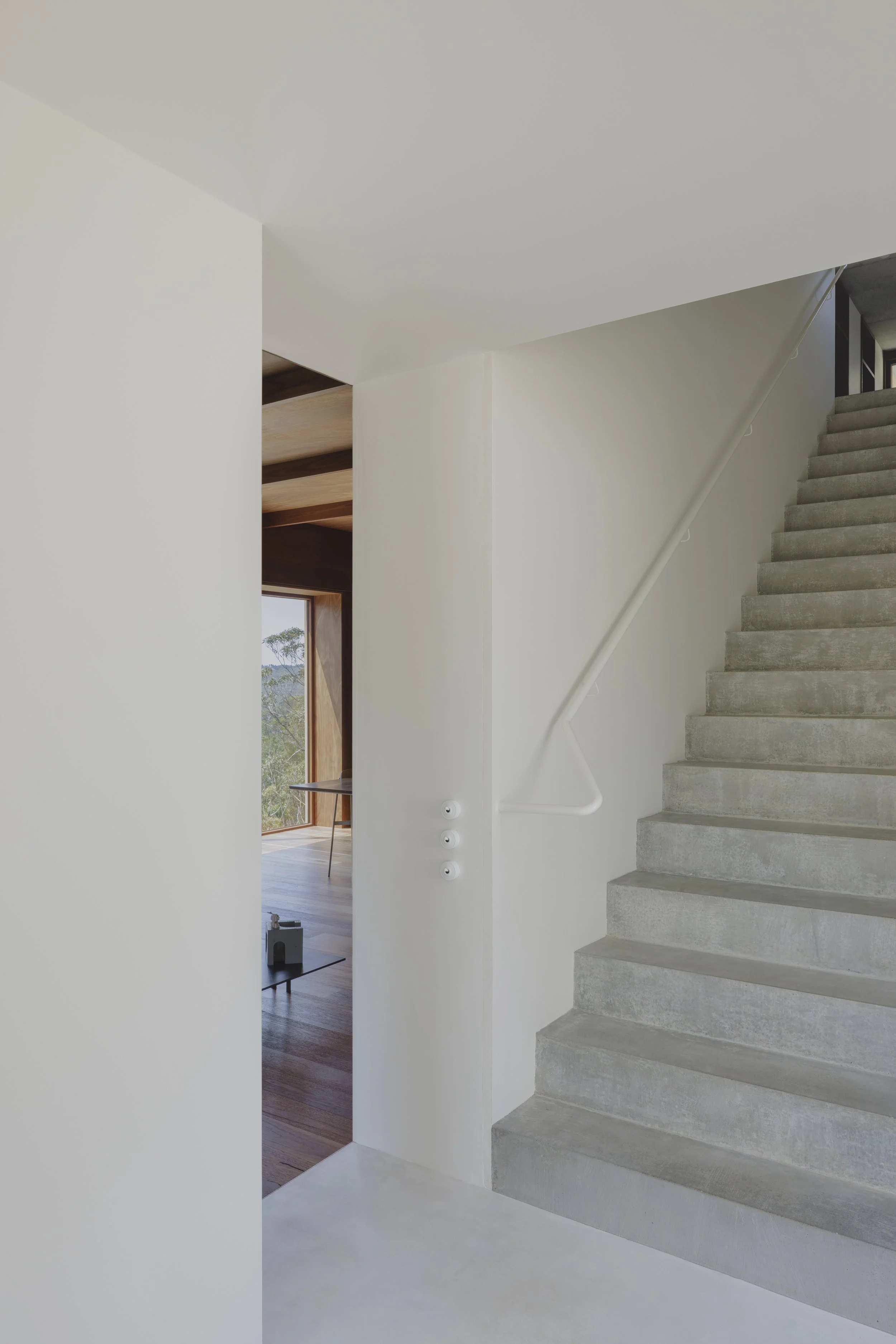 Interior view of a modern house showing a staircase with concrete steps, white walls, and a wooden floor visible in the adjacent room, with a window revealing trees outside.