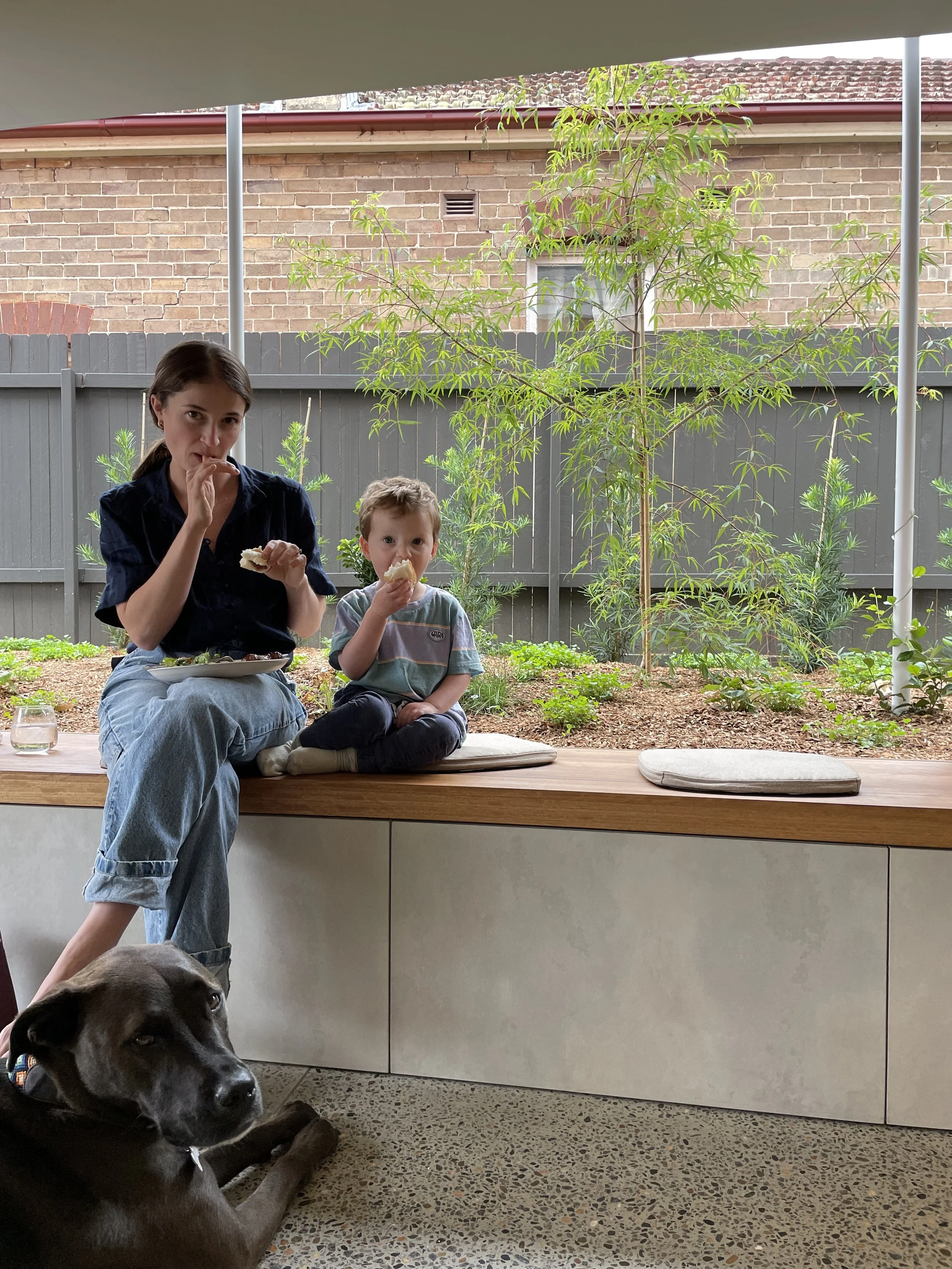 A woman and a young boy sitting on a wooden bench, eating bread. A dark-colored dog is lying on the floor nearby. Behind them is a garden with green plants and a small tree, enclosed by a gray fence and brick house.