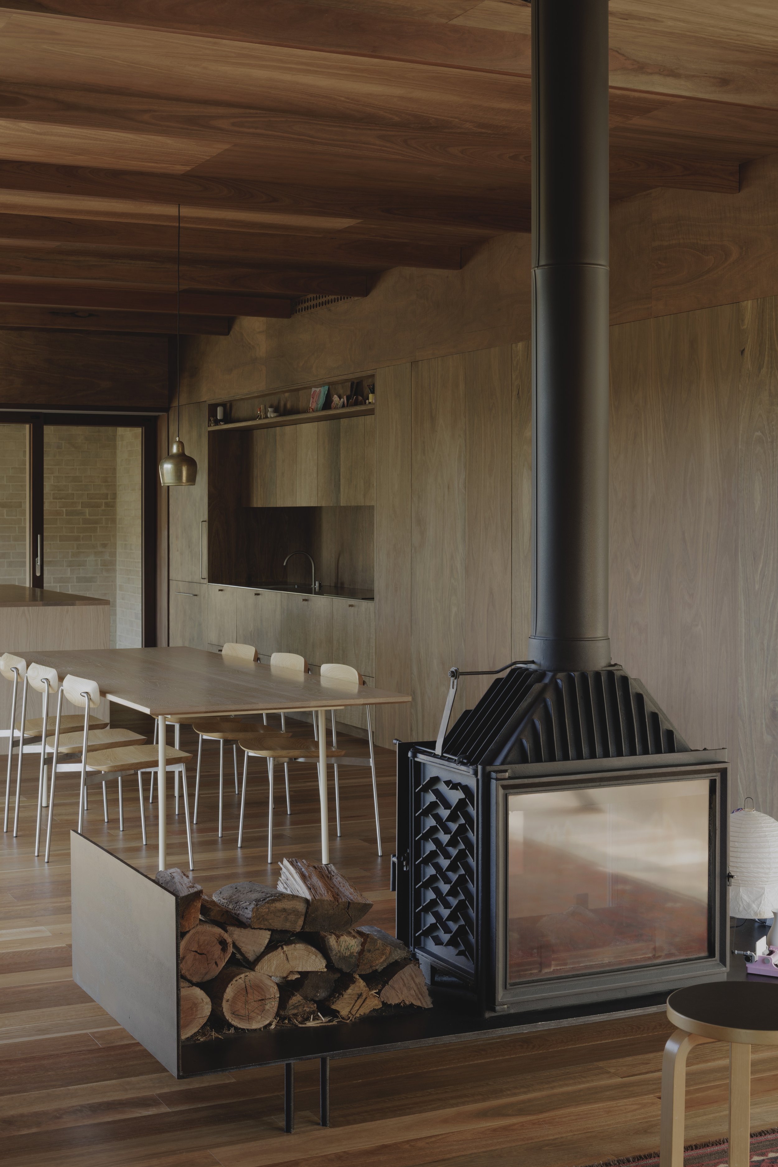 Interior view of a modern kitchen with wooden cabinets, a dining table with chairs, a wood-burning stove, and a section of wood logs stored in a metal holder, with wood flooring and a brick wall window in the background.