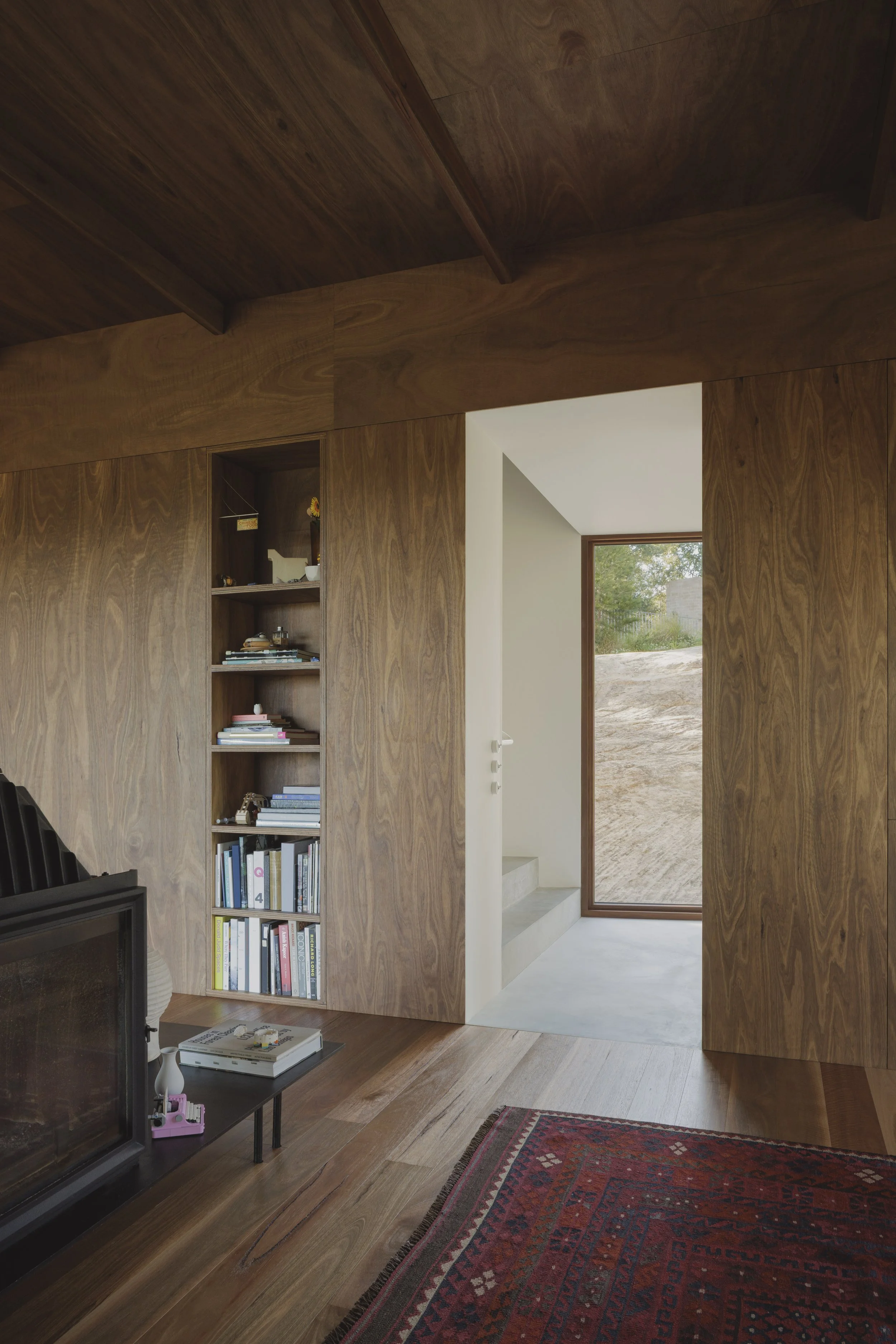 A room with wood-paneled walls and ceiling, a built-in bookshelf with books and decorative items, a dark fireplace, a red patterned rug, and an open doorway revealing an outdoor area with a window showing green trees and a grassy landscape.