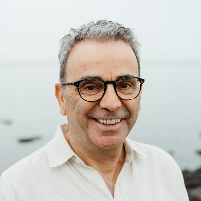 A middle-aged man with gray hair, black glasses, and a white shirt smiling outdoors by a body of water.
