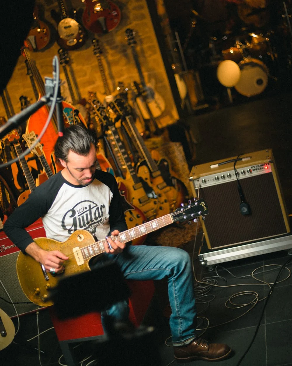 Nico Chona joue de la guitare électrique dans un lieu rassemblant de nombreuses guitares