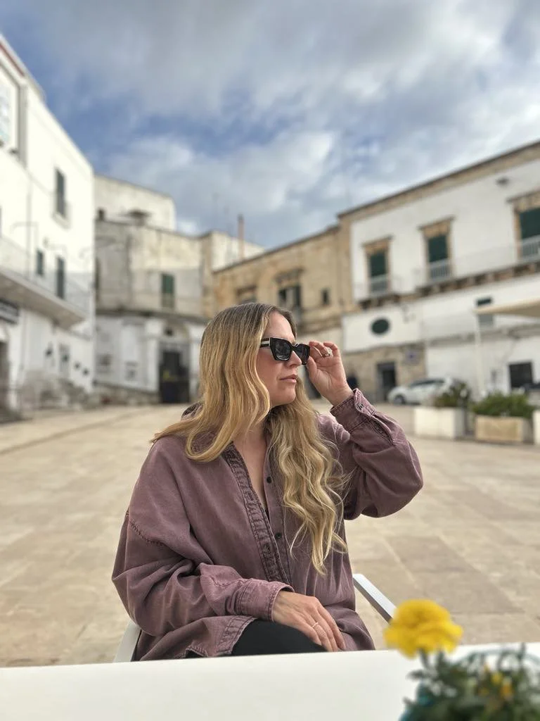 A woman with long wavy blonde hair wearing sunglasses and a mauve oversized shirt, sitting outdoors at a table with a blurred background of white buildings and a cloudy sky.