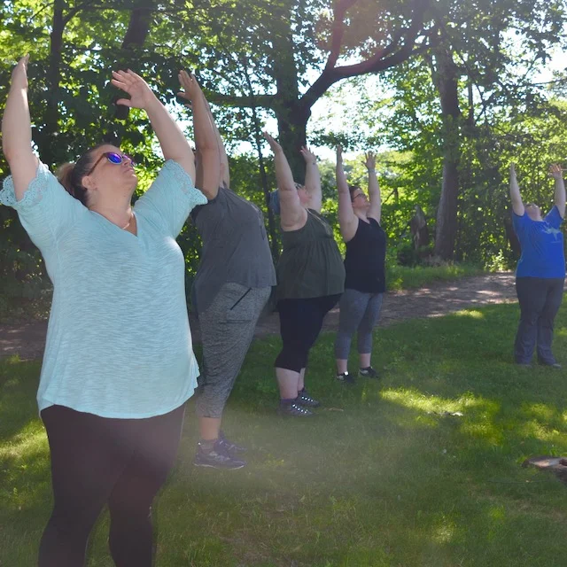 Image description: people with the arms raised overhead in a yoga pose, outside with greenery all around.