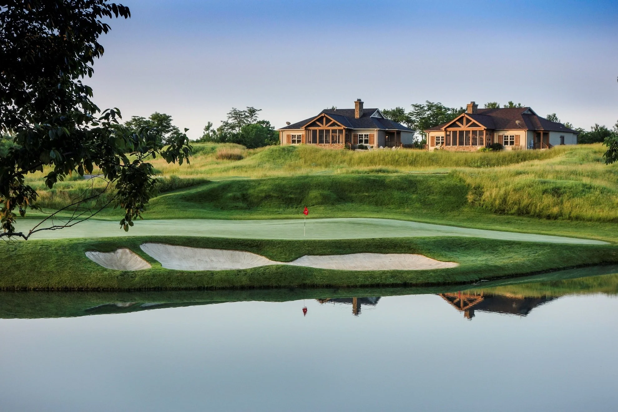 A pond in the foreground, a golf hole with a red flag in the middle, and two cottages in the background.