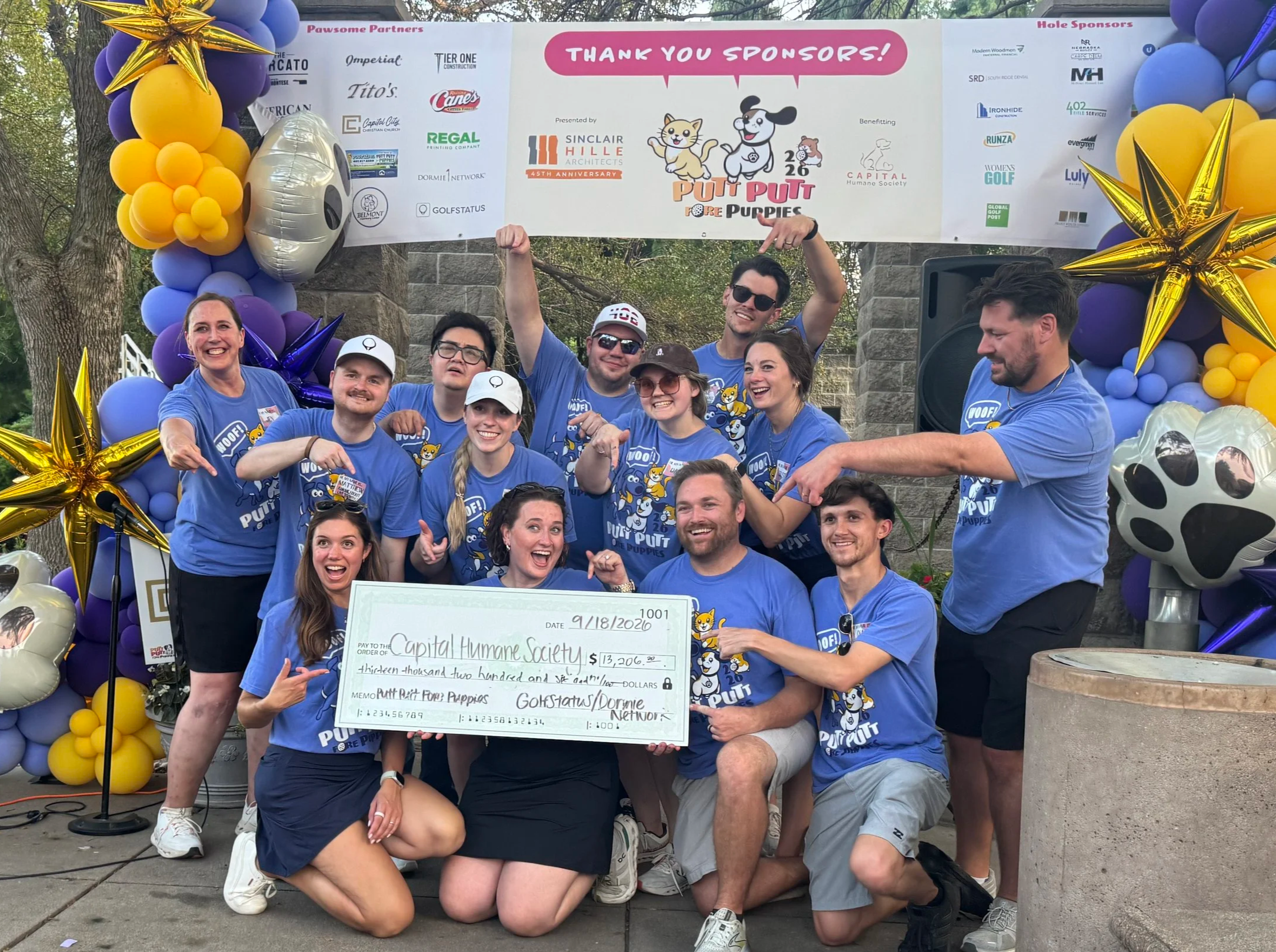 A golf tournament planning team in blue t-shirts points to a big check showing the proceeds from the event.