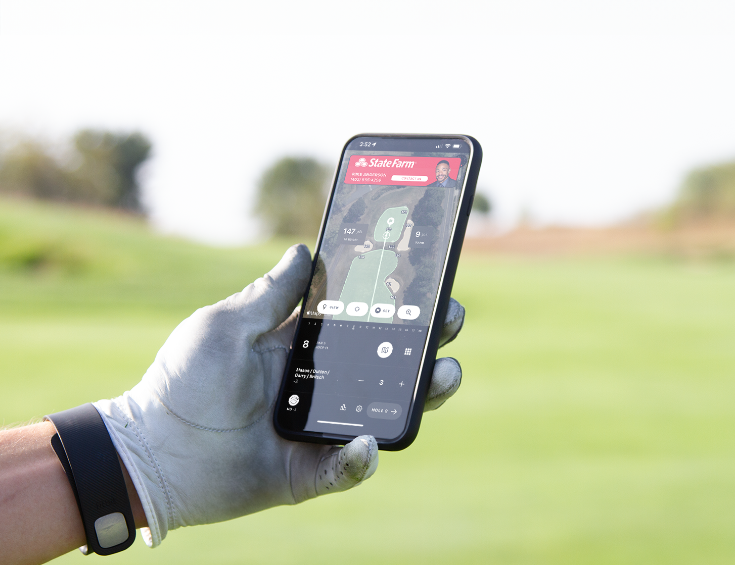 A golfer holds a smartphone showing a sponsor logo in a live scoring app.