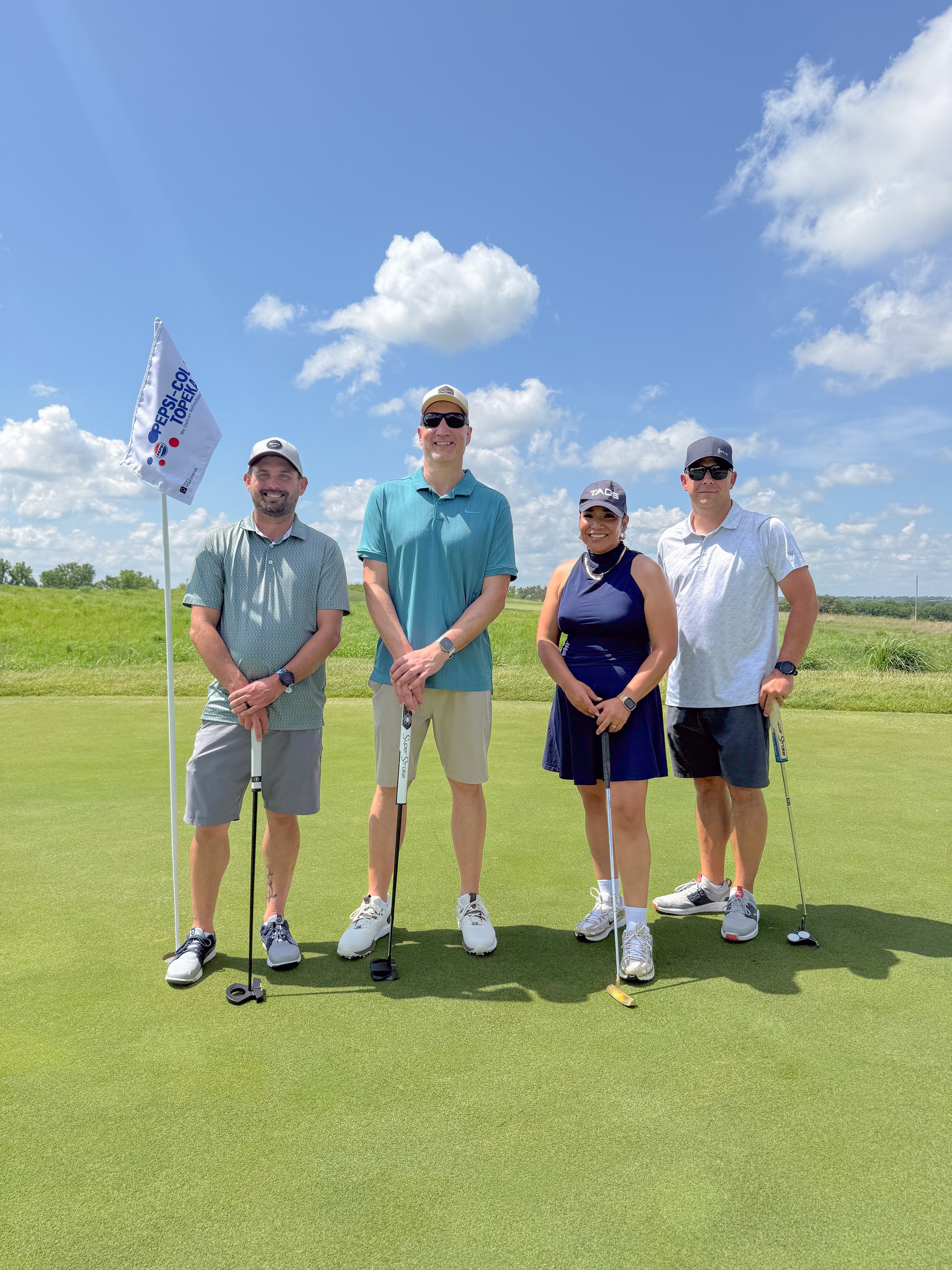 Four golfers stand on a green next to a custom pin flag at a charity golf tournament.