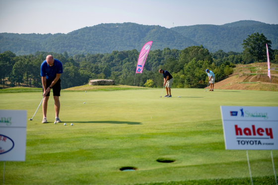 Three men practice their putts on the putting green, surrounded by sponsor signage at a golf fundraiser.