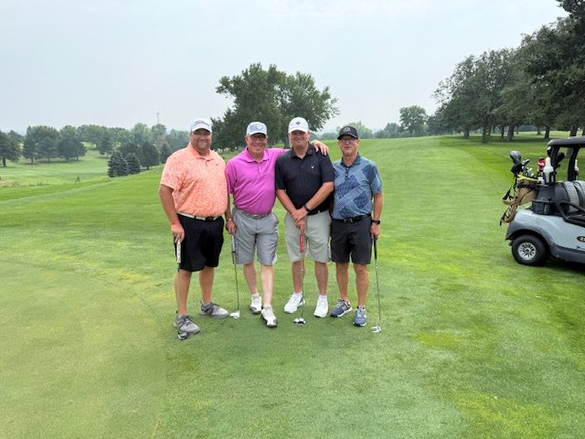 Four golfers pose on a golf course at a charity golf tournament.