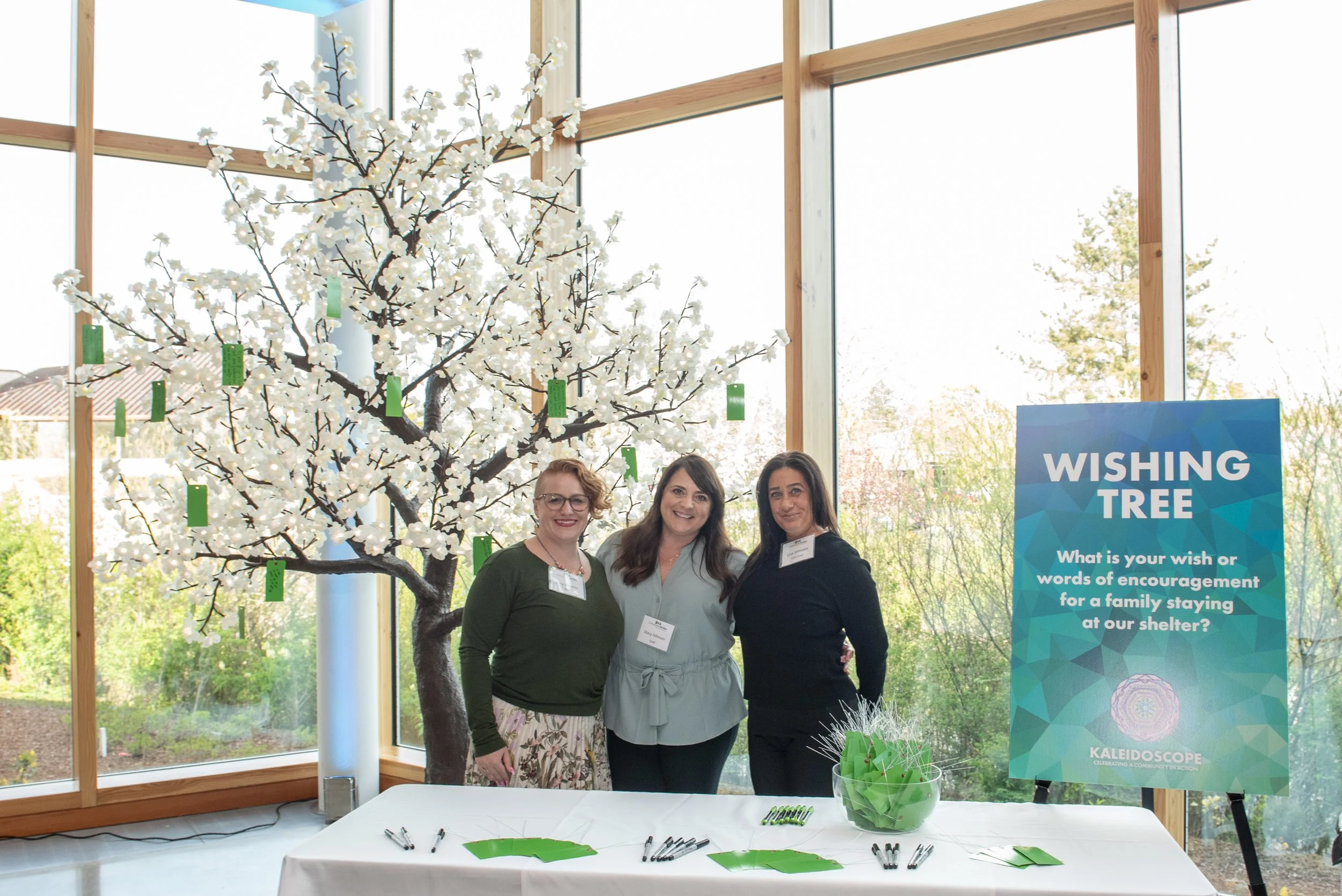 Three women pose behind a table at a fundraising event, inviting people to write words of encouragement.Three women pose behind a table at a fundraising event, inviting people to write words of encouragement.