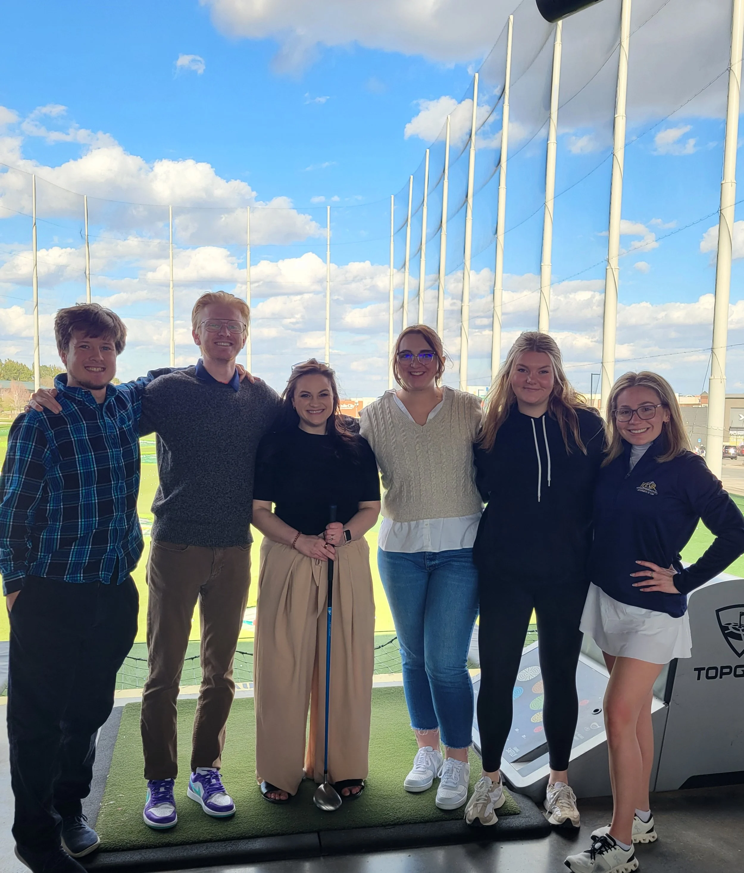 Two male and four female golfers pose with a golf club at a TopGolf fundraiser.