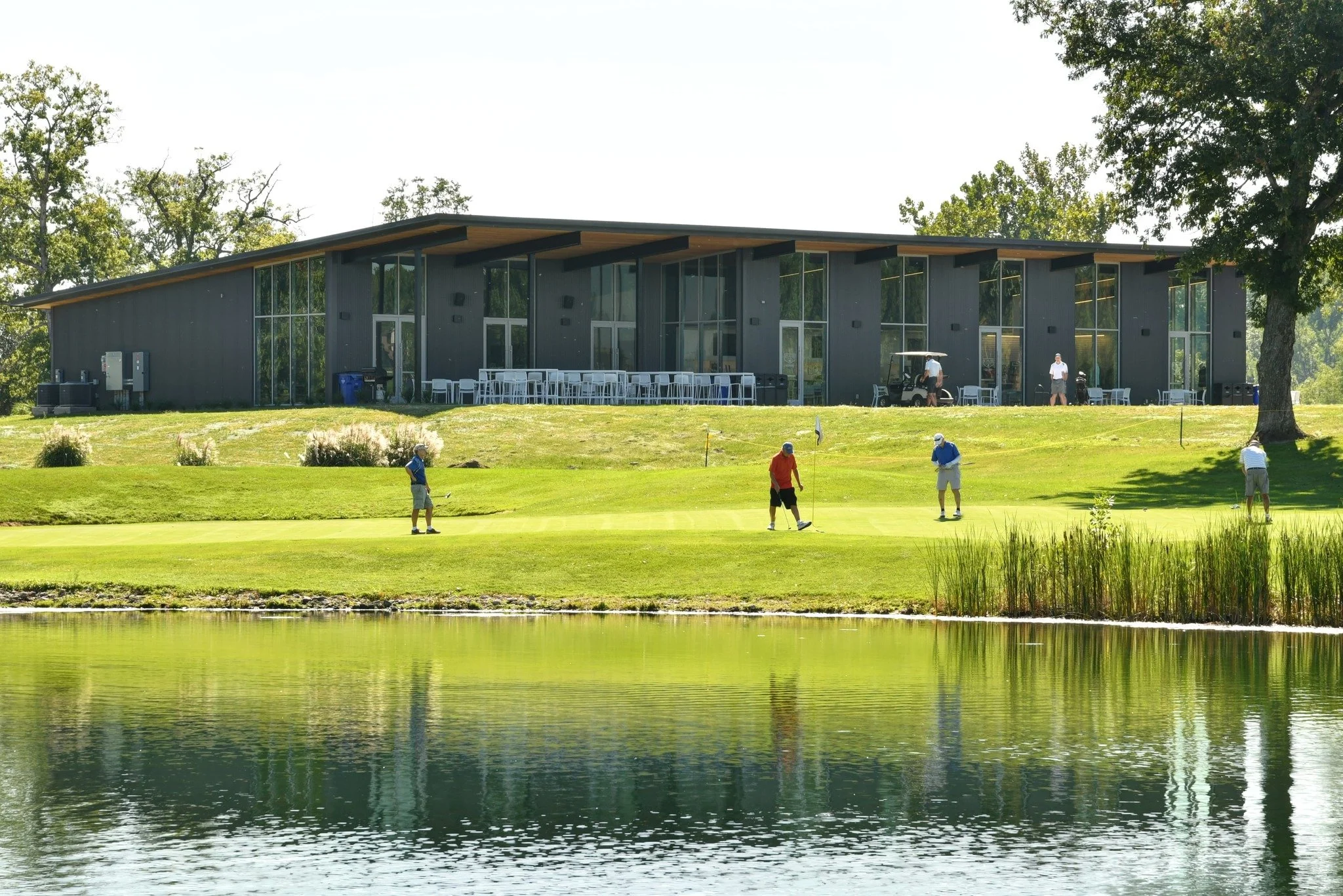 Four golfers putt on the practice green in front of the Ellis Golf Course Clubhouse, with a pond in the foreground.