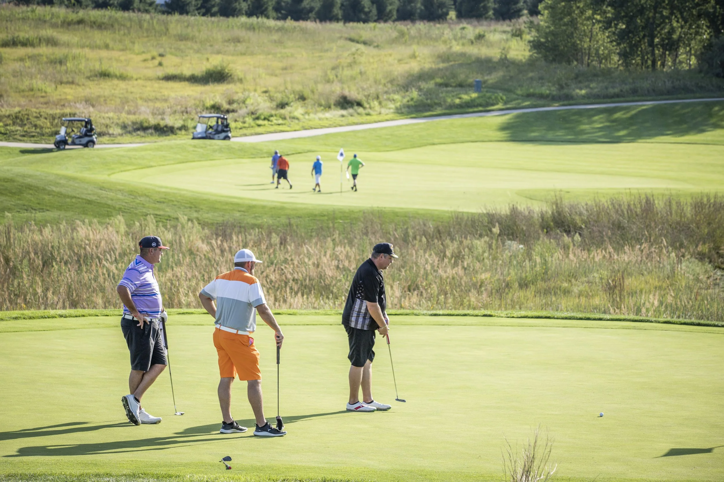 Three golfers on a green at a golf course in the foreground, with four golfers in the background.