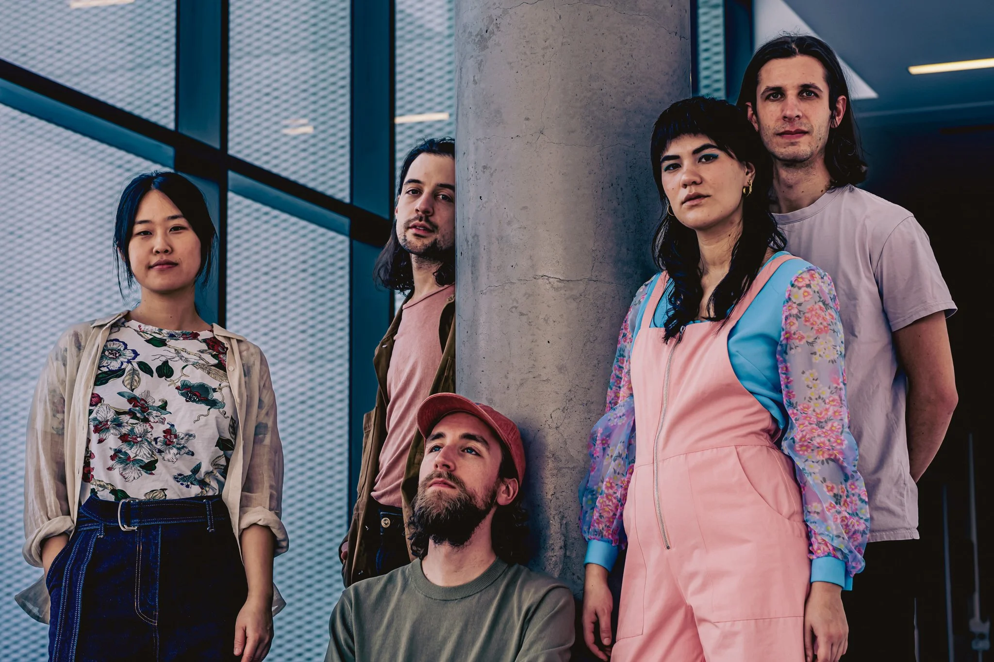 Band members of Pony Girl stand around a cement pillar in a modern building in front of large windows. They are looking into the camera, and wearing casual clothing in soft pink, blue, and neutral hues.