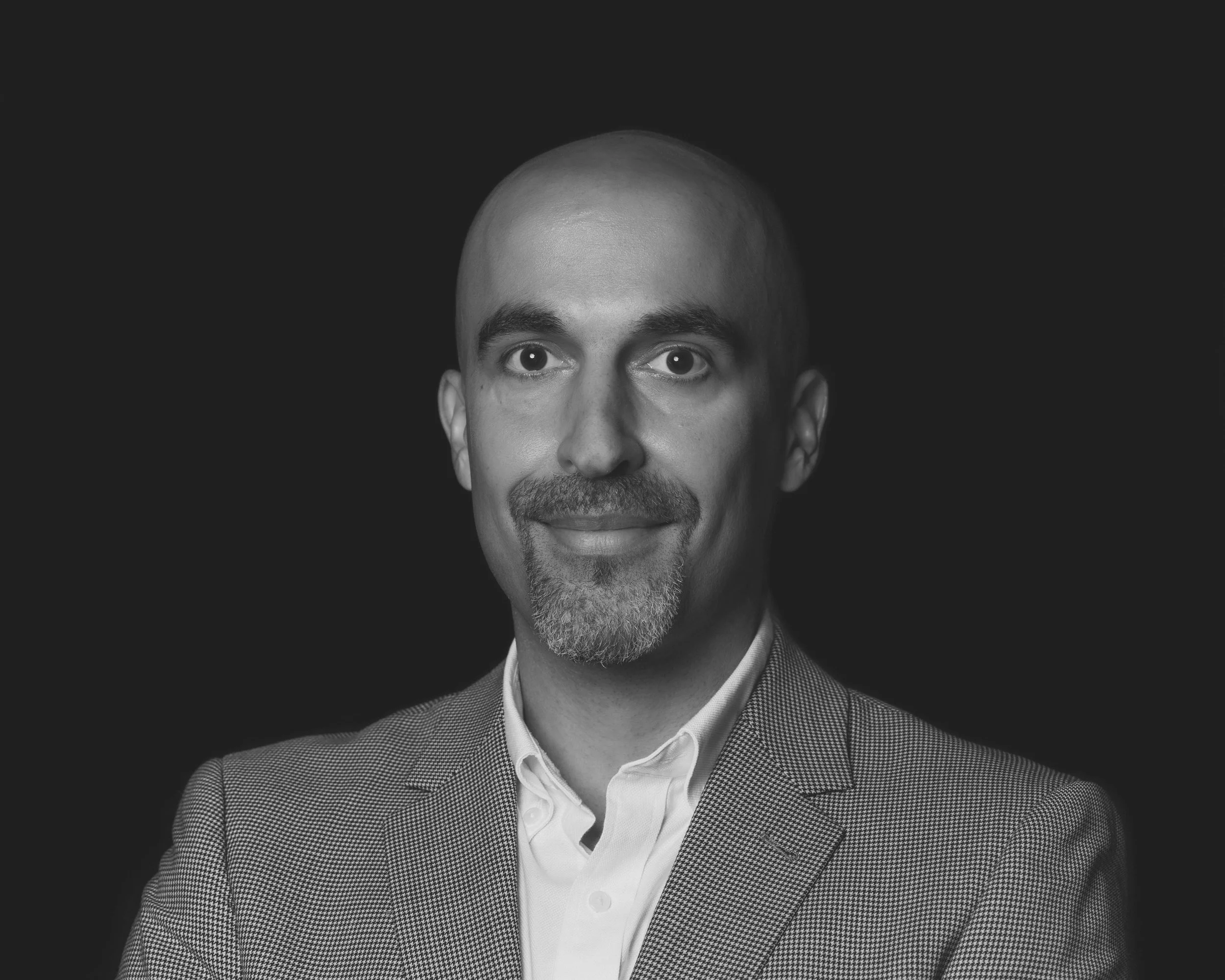 Black and white photo ofAmin Alsadenstanding in front of a black background wearing a white button up shirt and a grey suit jacket. Amin has a slight smile.