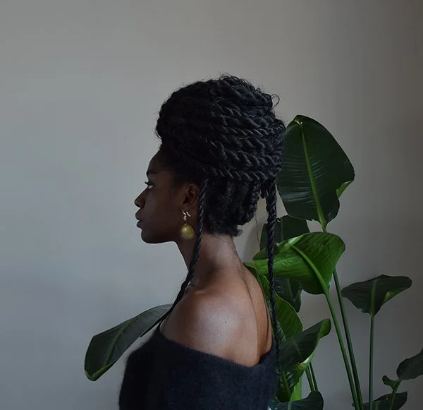 Kosisochukwu sits in profile facing the back corner of the naturally-lit room. She wears a black sweater that sits off her shoulders, and a gold earring hangs from her ear. A large plant sits behind her.