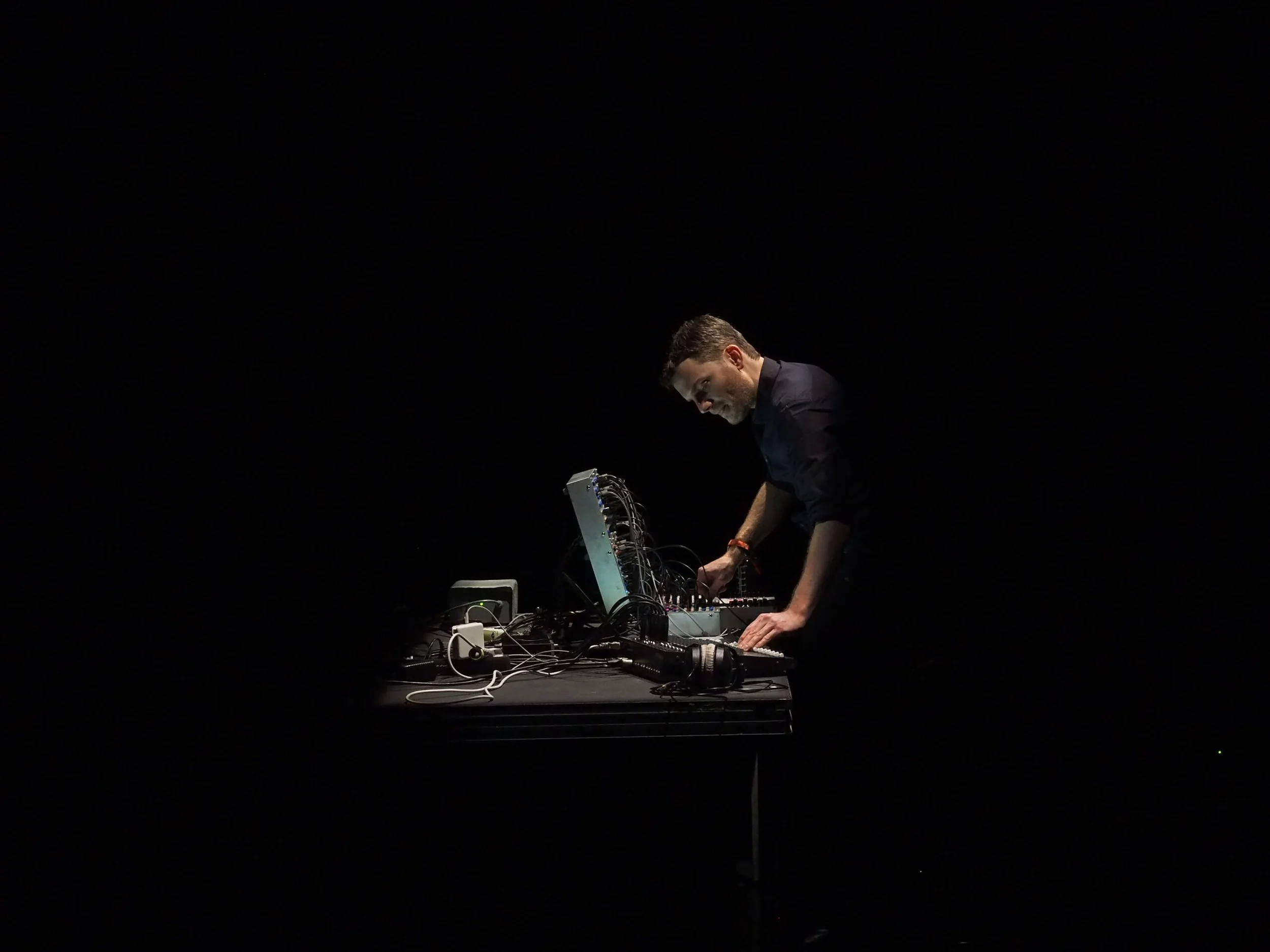 Thomas Ankersmit leans over a table in an entirely darkened space. On the table is a modular synthesizer, headphones, and various cables. He is wearing a black collared shirt.