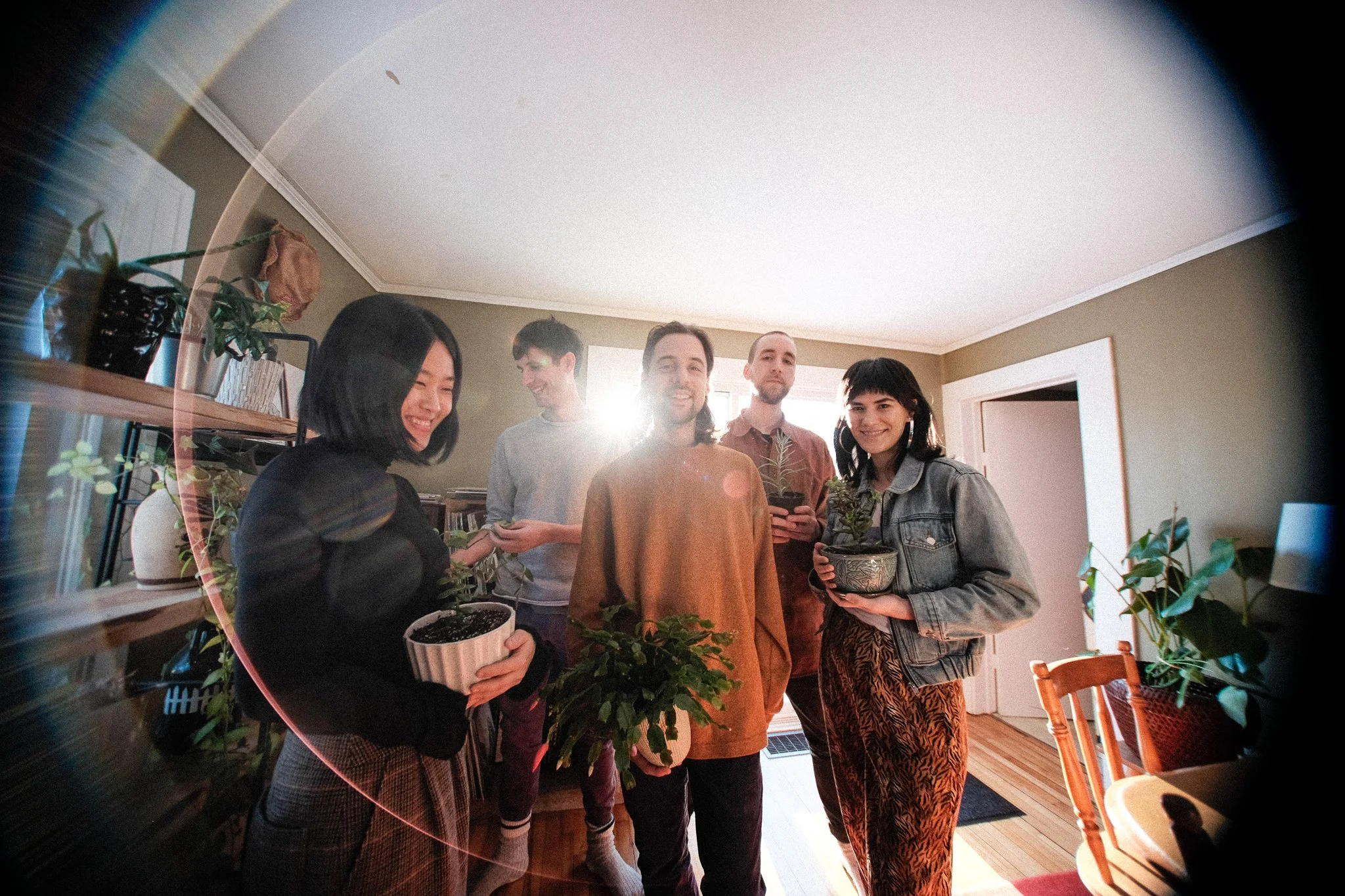 The band members of Pony Girl are standing in an apartment holding house plants. Sunlight streams through a back window, and the band members are smiling.