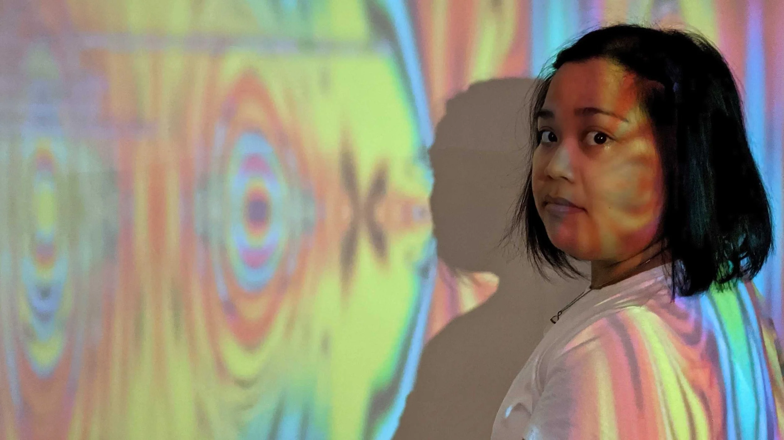 A Filipinx woman with black shoulder length hair wearing a white shirt and standing in front of a colourful projection