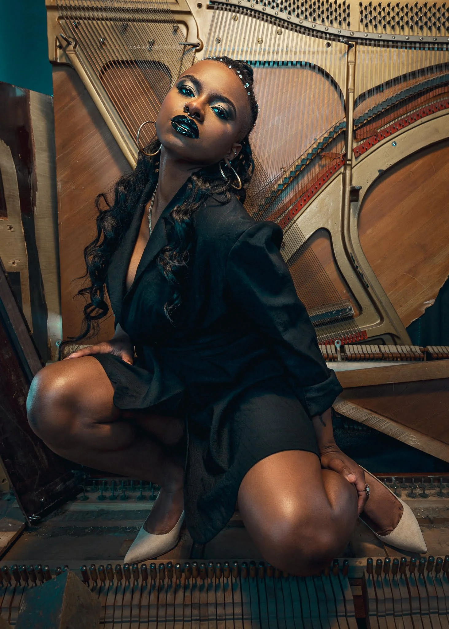 Morgan-Paige crouches on and in front of a piano's sound board. She is wearing a black dress, white heels, black lipstick, and hoop earrings.