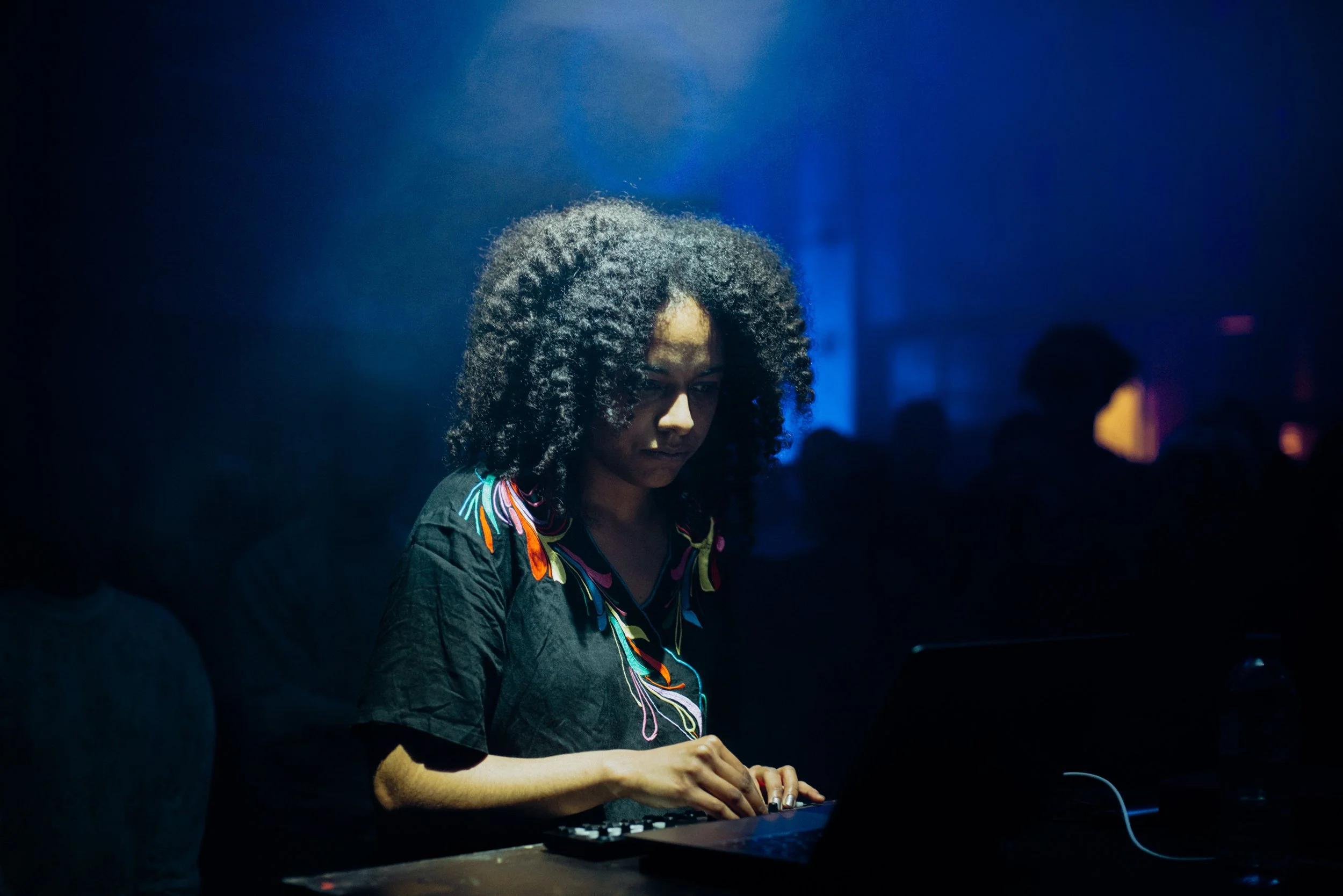 Jessica Ekomane stares off camera while manipulating a tabletop electronic instrument. She is in a dark room with an audience listening behind her. She is wearing a black shirt with colorful embroidery.