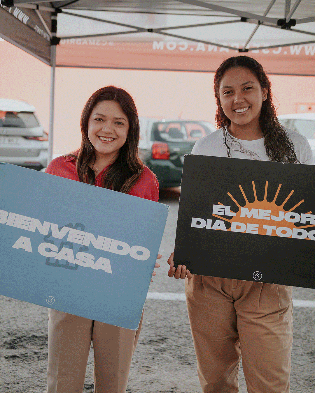 Dos mujeres sosteniendo carteles de celebración en un evento al aire libre, bajo toldo, con autos en el fondo.
