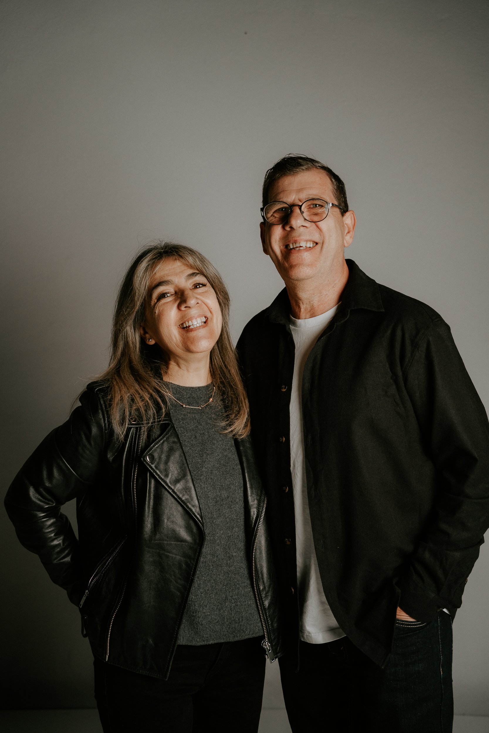 Una mujer y un hombre sonriendo, posando juntos contra un fondo gris, la mujer lleva chaqueta de cuero negra y el hombre lleva camiseta blanca y chaqueta negra.