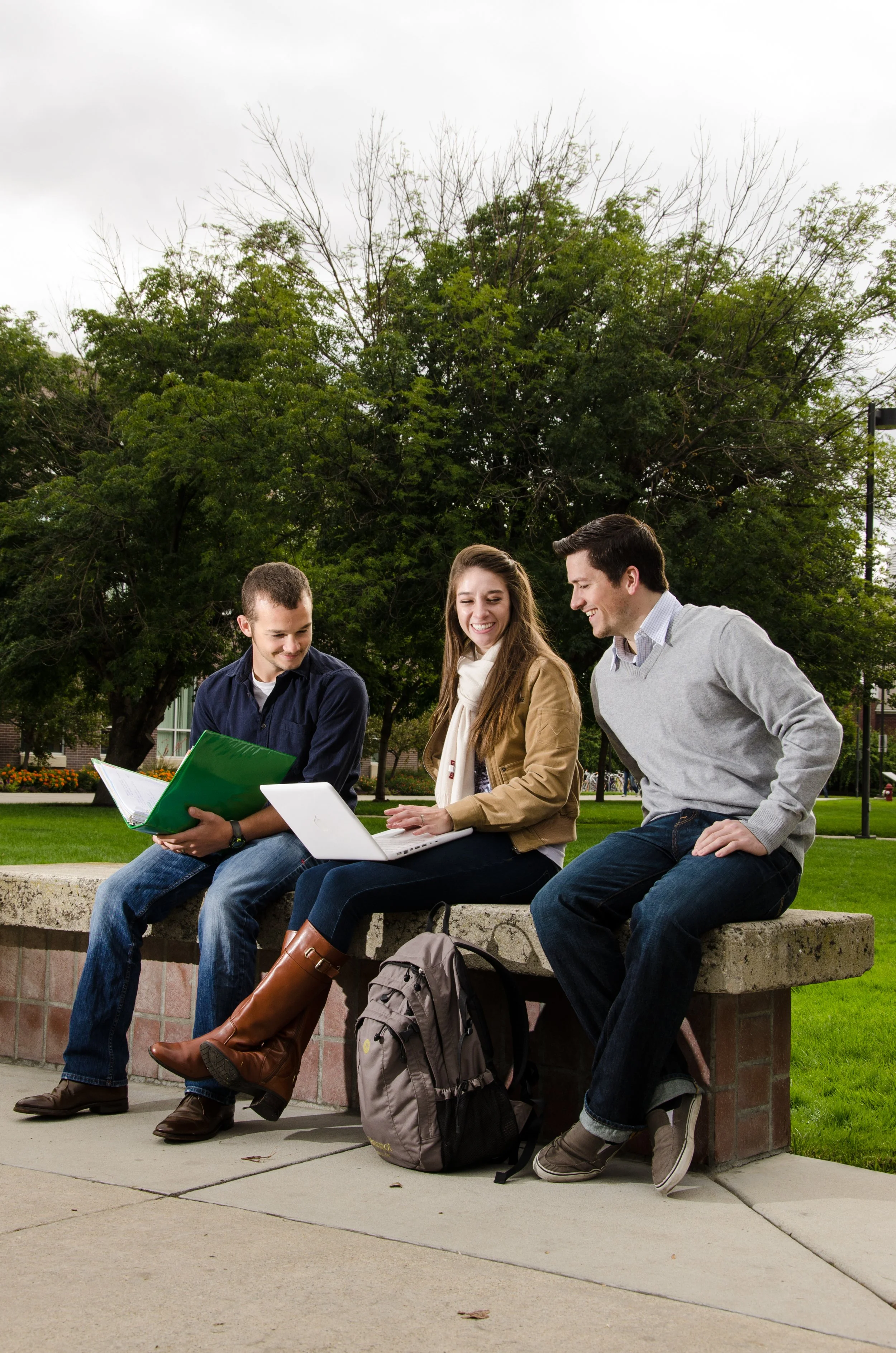 Three students sitting on a low brick wall outdoors, two men and one woman, looking at a laptop and having a conversation, with backpacks on the ground in front of them, and a large tree and cloudy sky in the background.