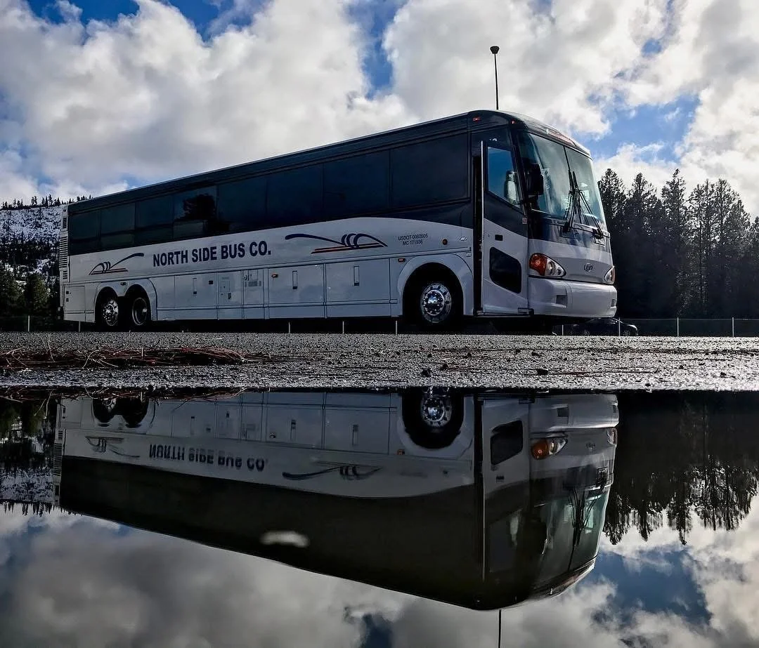 A gray and black bus with 'North Side Bus Co.' written on the side, parked on a gravel surface. Its reflection can be seen in a body of water below. There are trees and a partly cloudy sky in the background.