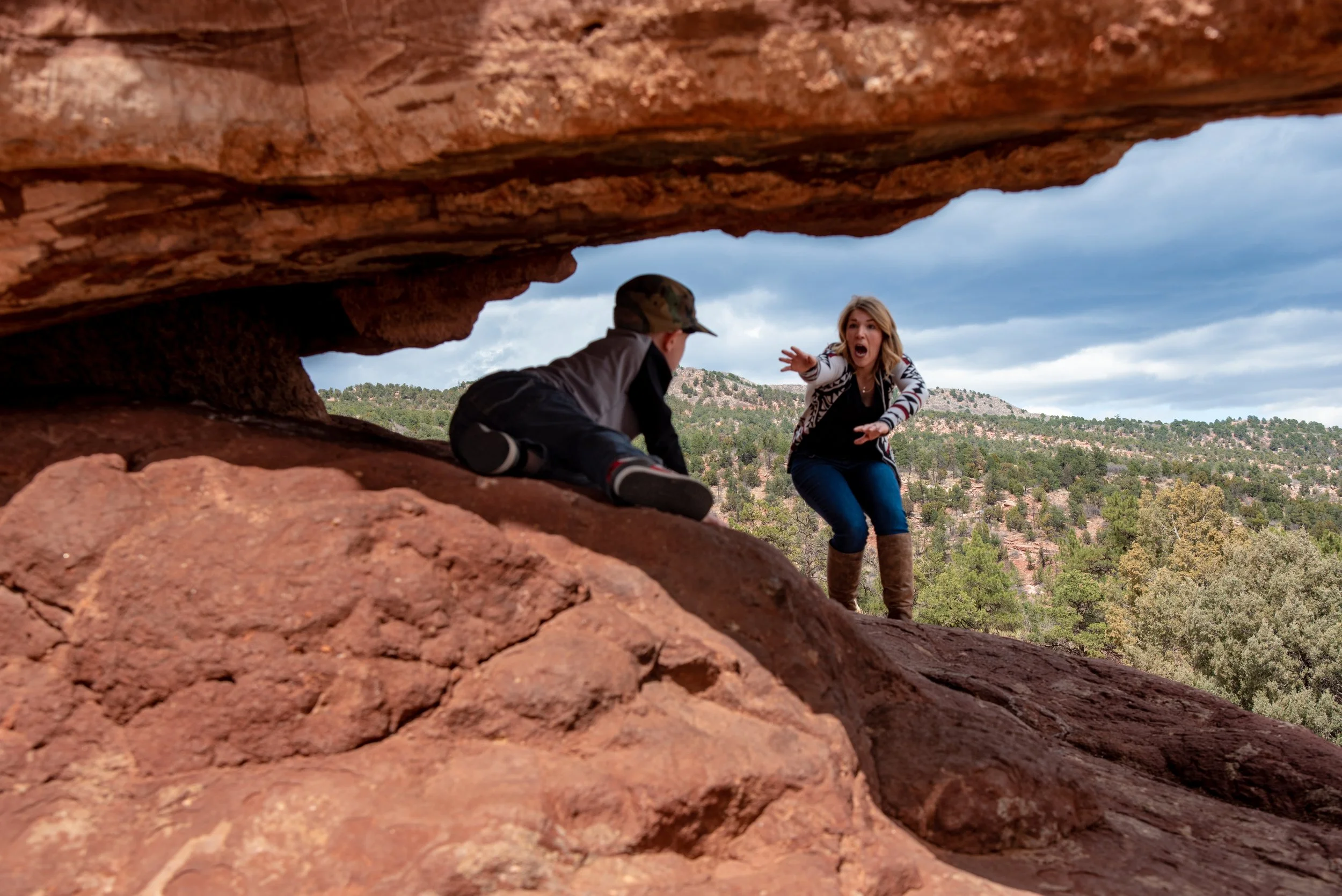 A woman reaching out to a boy lying on the ground under an orange rock formation with a scenic landscape of trees and hills in the background.