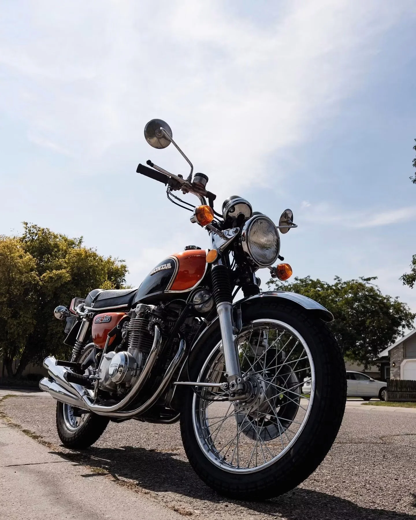 A vintage Honda motorcycle parked on a driveway with trees and houses in the background on a partly cloudy day.