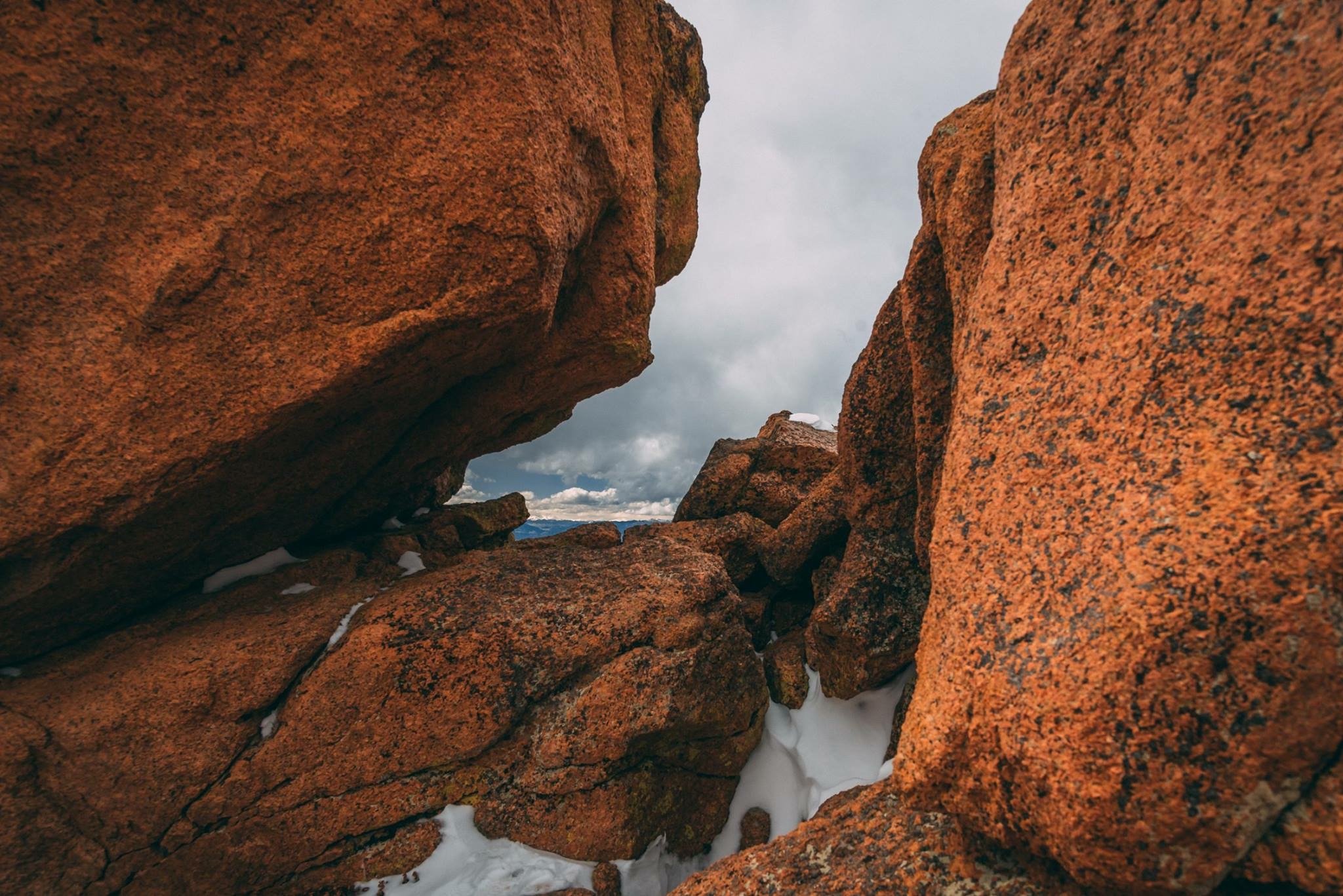 Large reddish-brown rocks with a gap between them, through which a cloudy sky and mountain range are visible in the distance, with patches of snow on the rocks and ground.