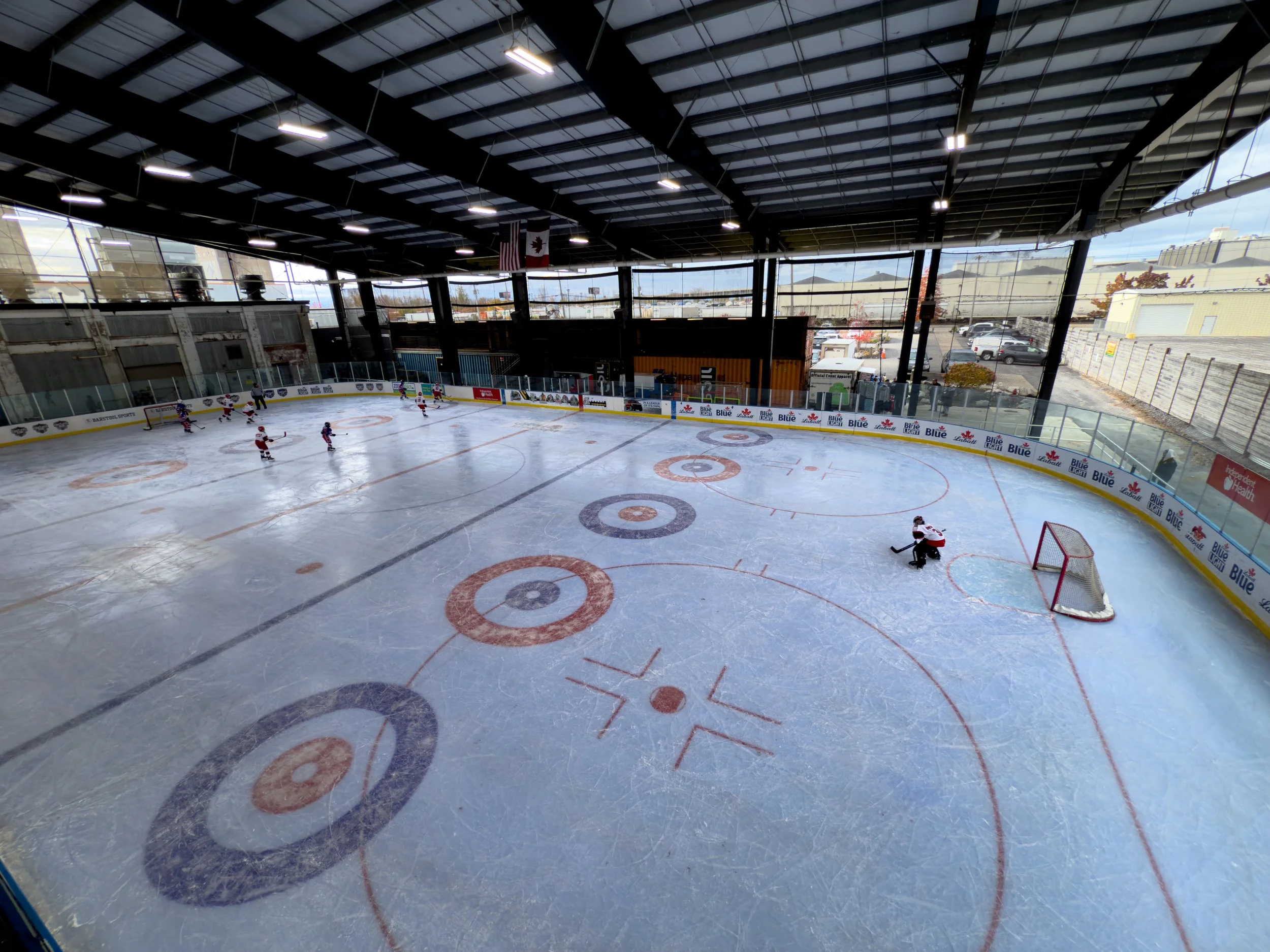Indoor ice hockey rink with players practicing on the ice, surrounded by clear protective barriers and advertising banners, with a view of the outside parking lot through large windows.