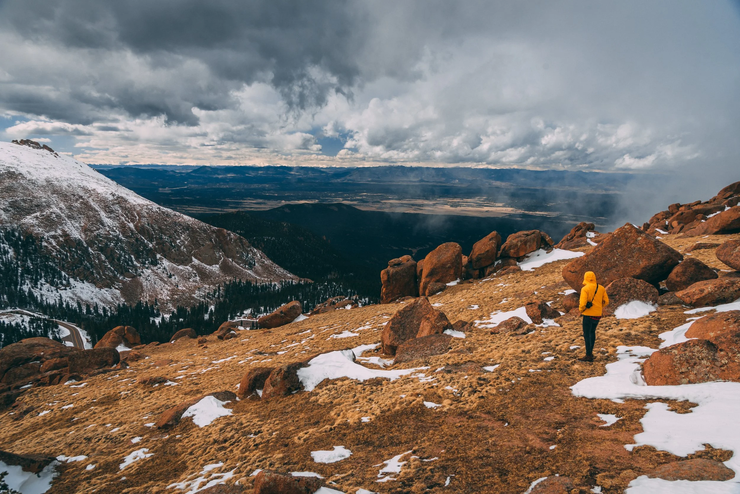 Hiking trail on a mountain with snow patches, rocky terrain, and a person in a yellow jacket looking at the landscape under cloudy skies.