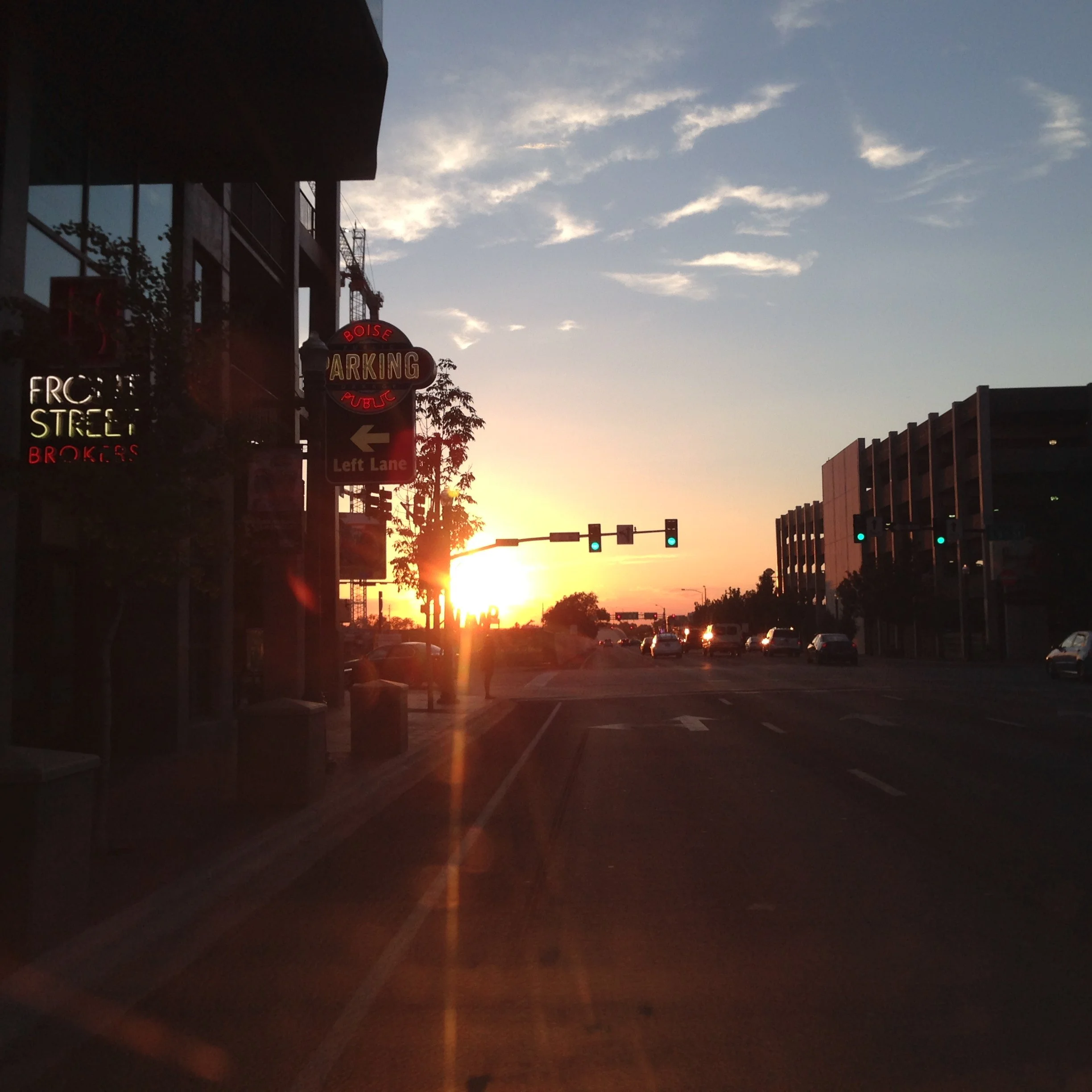 A city street during sunset with vehicles, modern buildings, and traffic lights, a sign indicating parking and directions, and a partly cloudy sky.