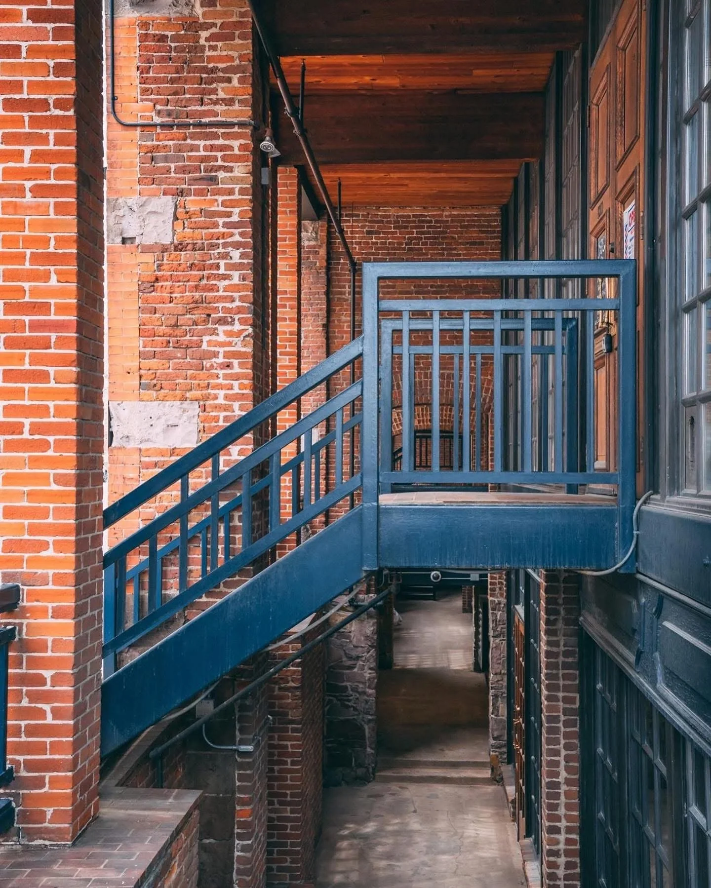 A multi-story brick building with exterior metal staircases and walkways, seen from a narrow walkway.