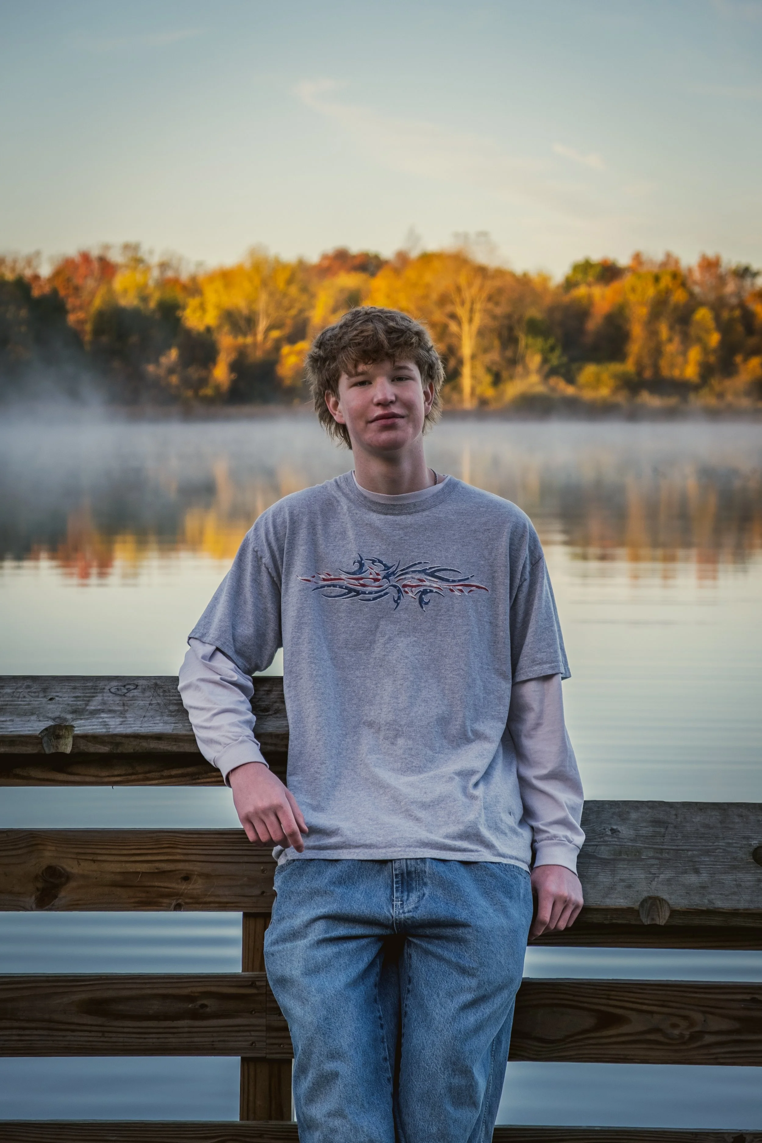 A teenage boy standing by a wooden railing near a body of water with a misty surface, with trees displaying fall foliage in the background during sunrise or sunset.