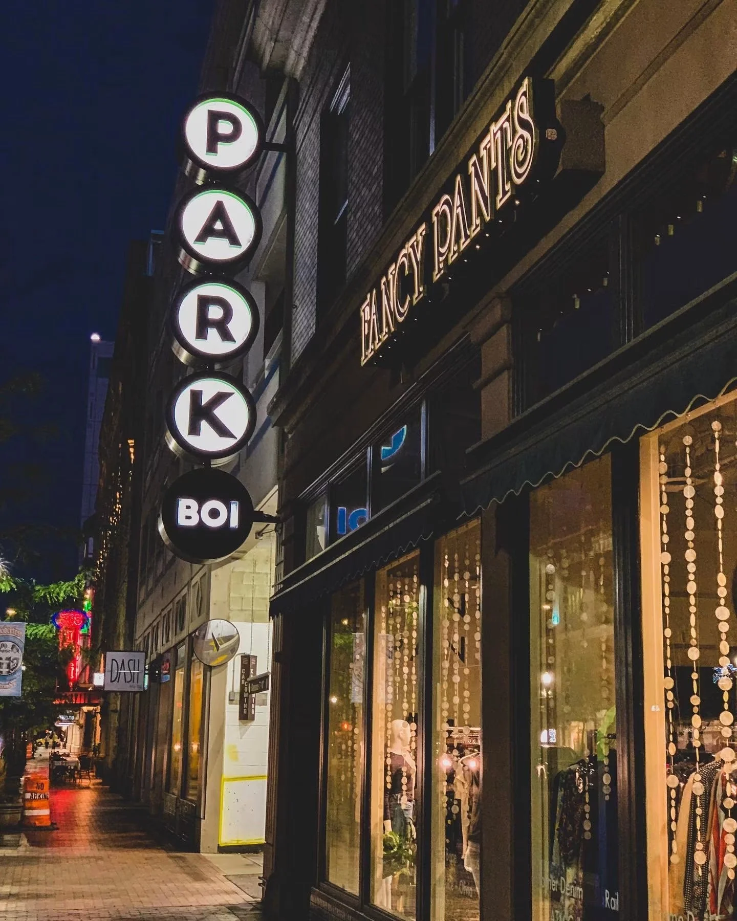 Nighttime street view with illuminated signs reading 'PARK' vertically and 'BANCY PANTIES,' along with storefront windows decorated with lights and displays.