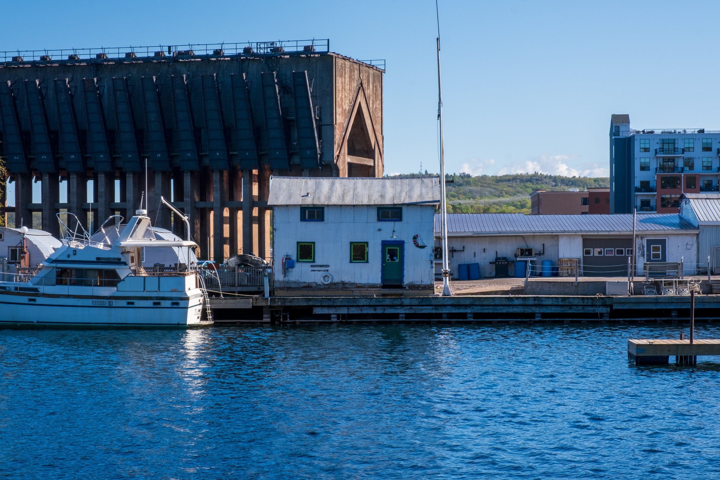 A marina with a white yacht docked in the water, surrounded by buildings including a large wooden structure, a small white house, and a modern blue building, with hills and clear blue sky in the background.