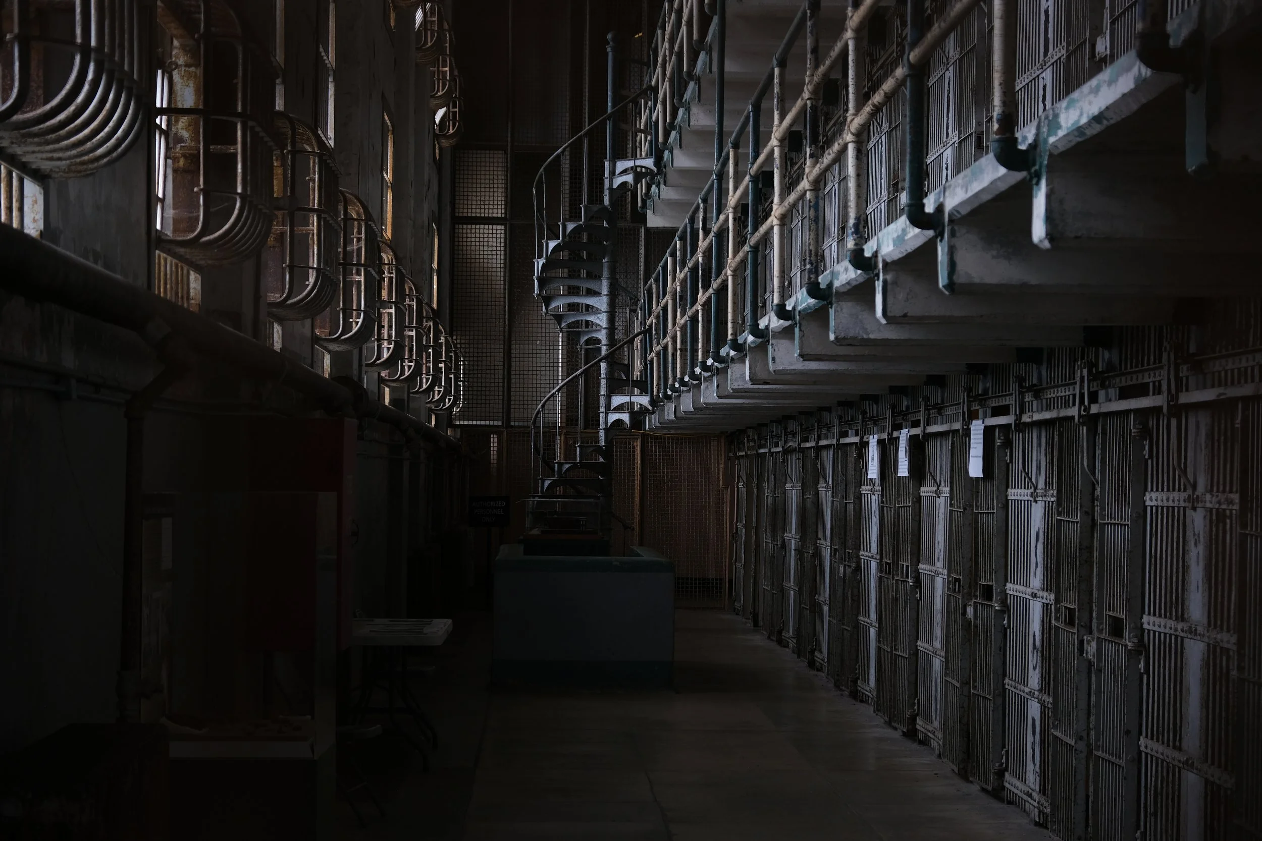 Image of an industrial or mechanical area with metal shelves or crates stacked along the walls, metal pipes running along the ceiling, and a spiral staircase in the center, all in a dark, shadowy setting.