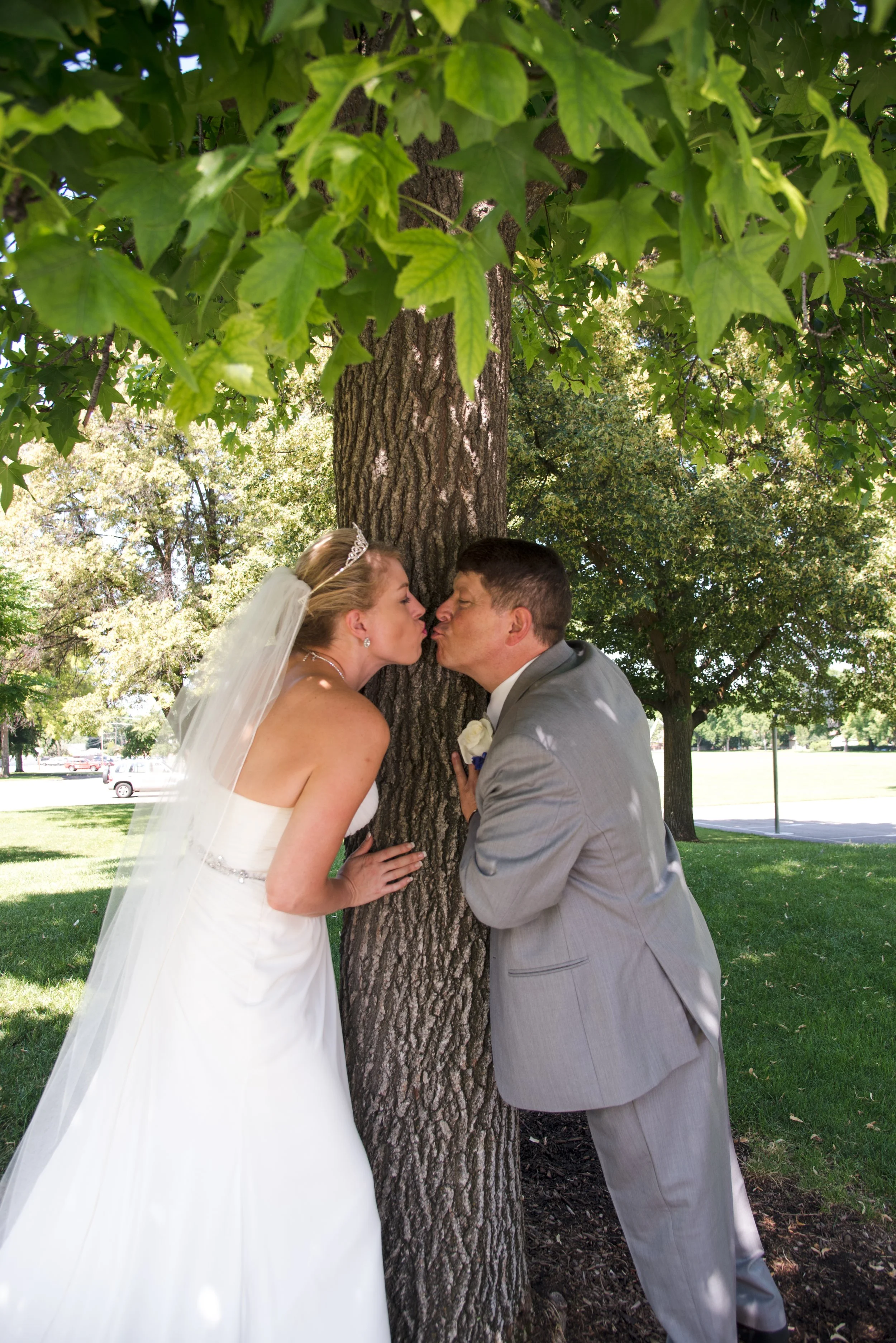 A bride and groom kissing behind a tree in a park on their wedding day, with green leaves and trees in the background.
