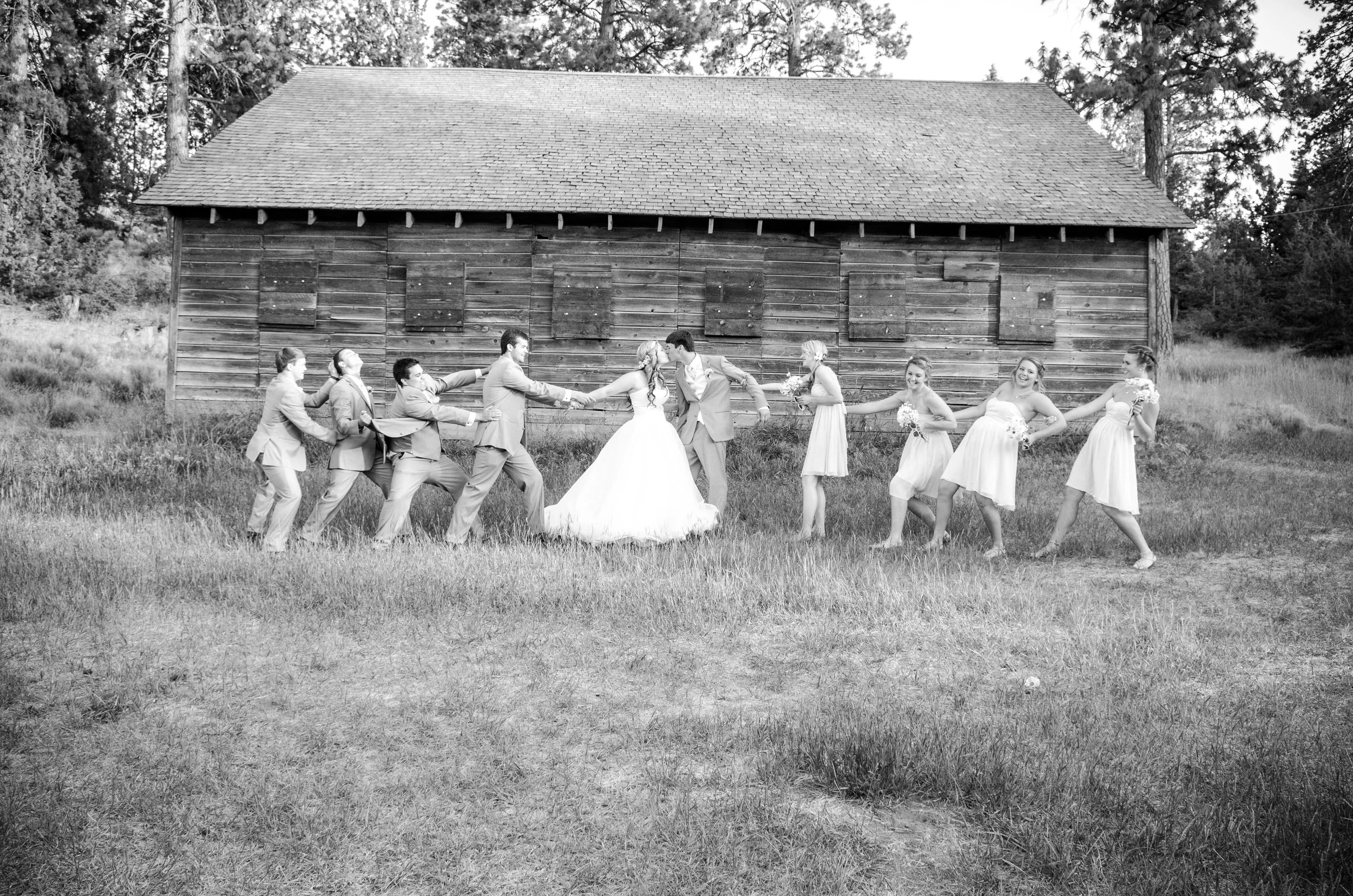 Black and white photo of a wedding party pulling on a rope in a field in front of a wooden barn. The bride and groom are in the center, holding hands, with other wedding guests, including bridesmaids and groomsmen, pulling on either side.