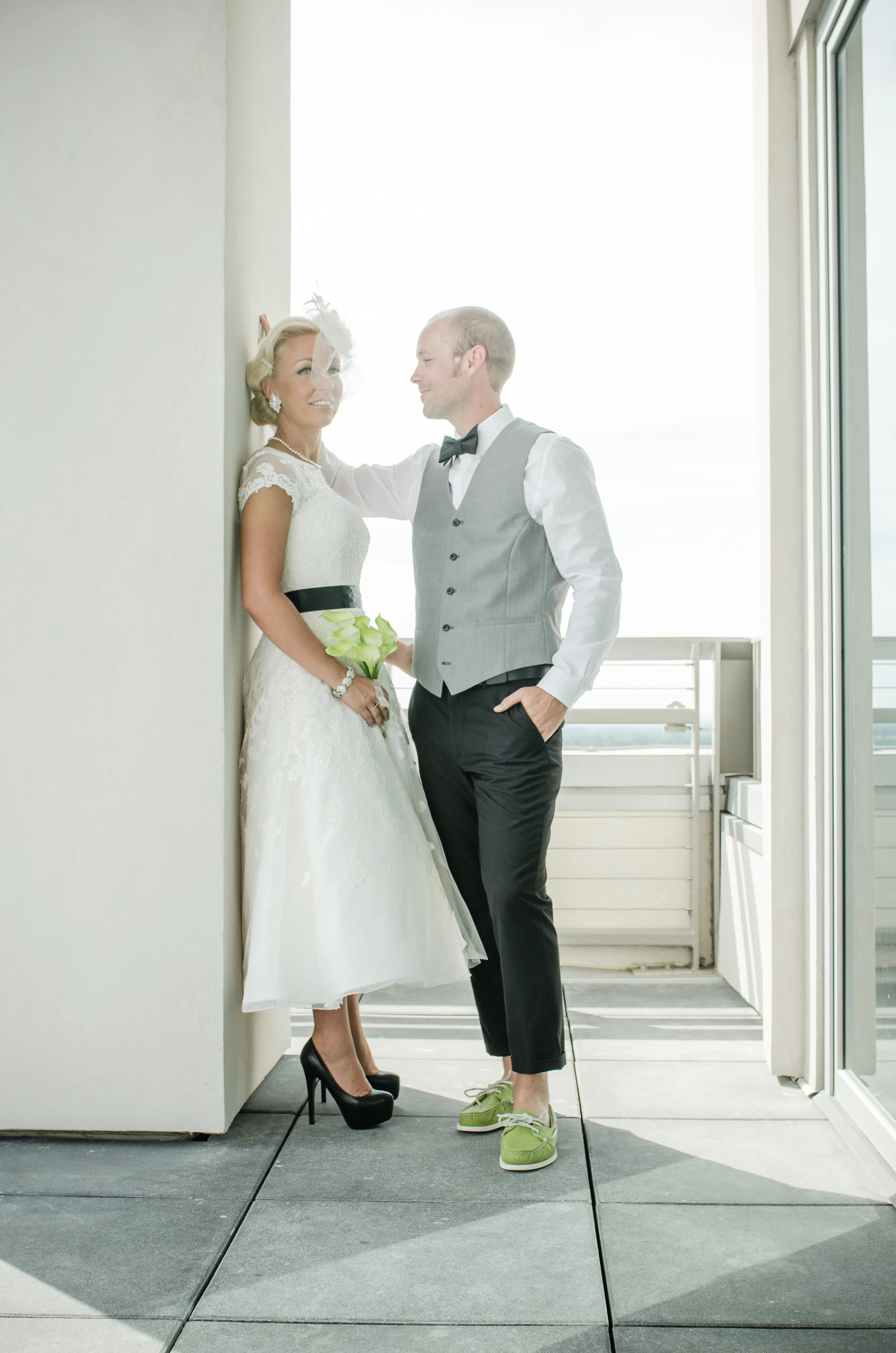 A bride and groom are posing together by an open door. The bride is dressed in a white wedding gown with lace details, wearing black high heels, pearl jewelry, and holding a bouquet of calla lilies. The groom is wearing a light grey vest, black trous