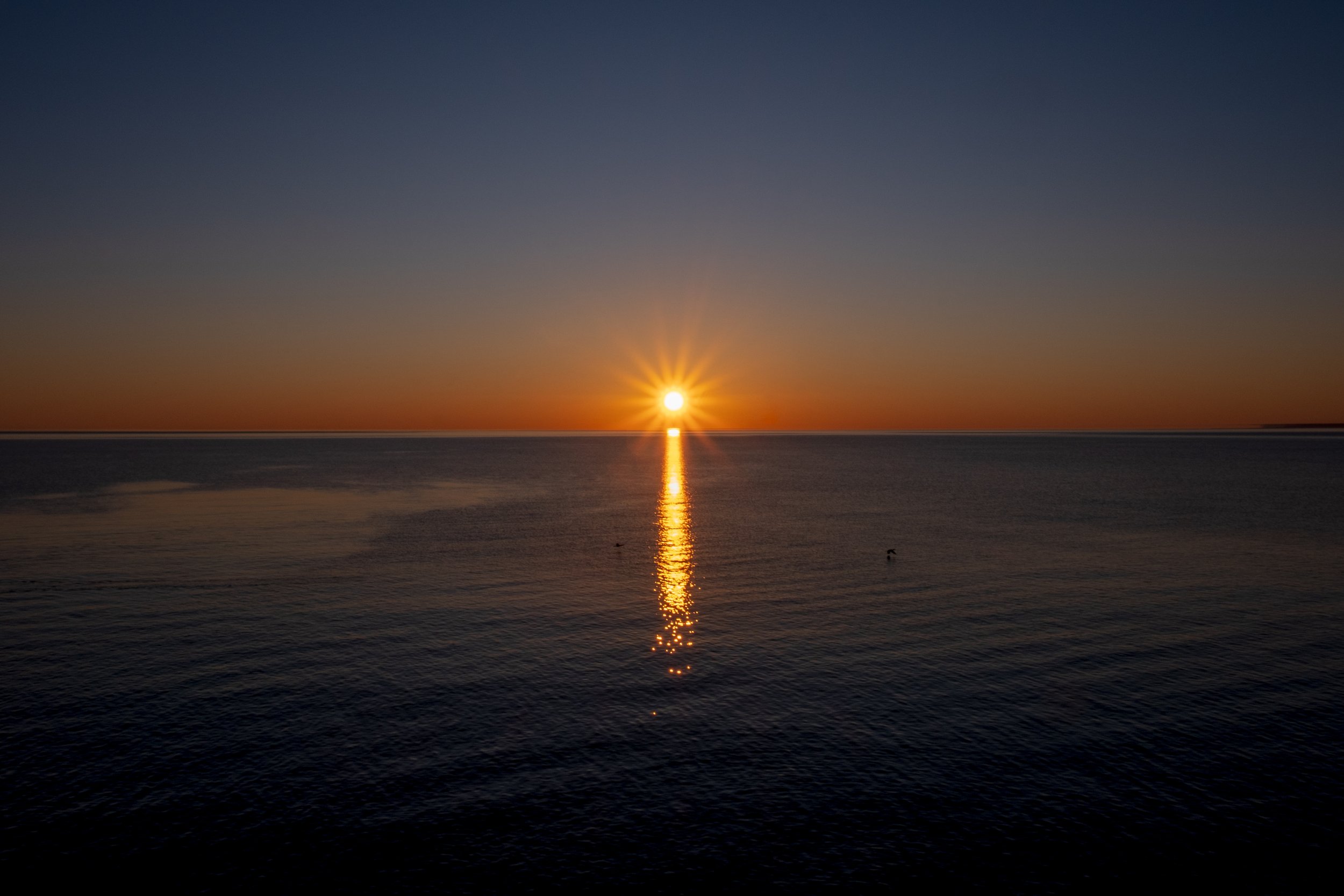 Sunset over a calm body of water, with the sun near the horizon and its reflection on the water surface.