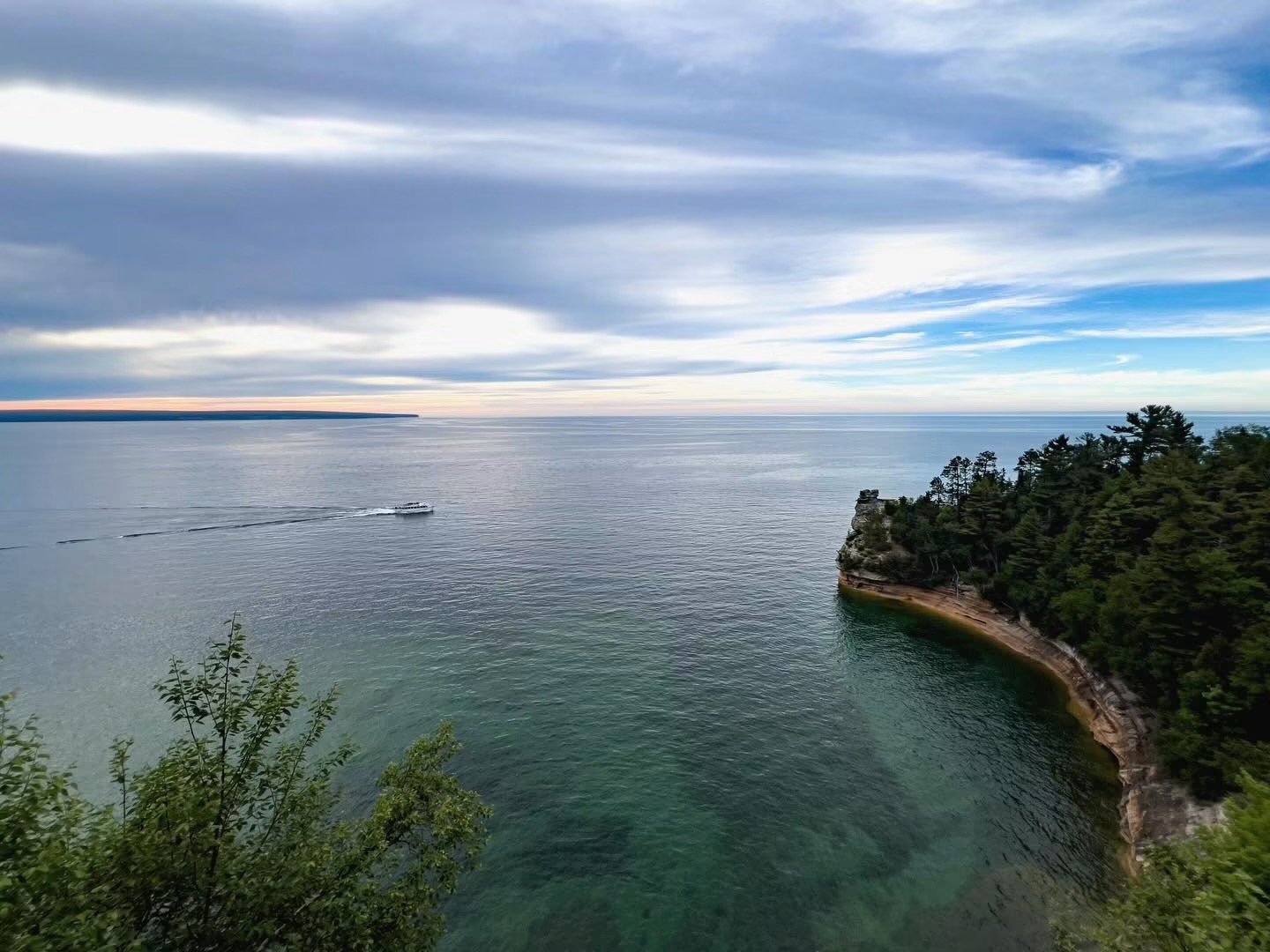 Scenic view of a lakeshore with a boat leaving a wake, a forested cliff on the right, and a cloudy sky above.