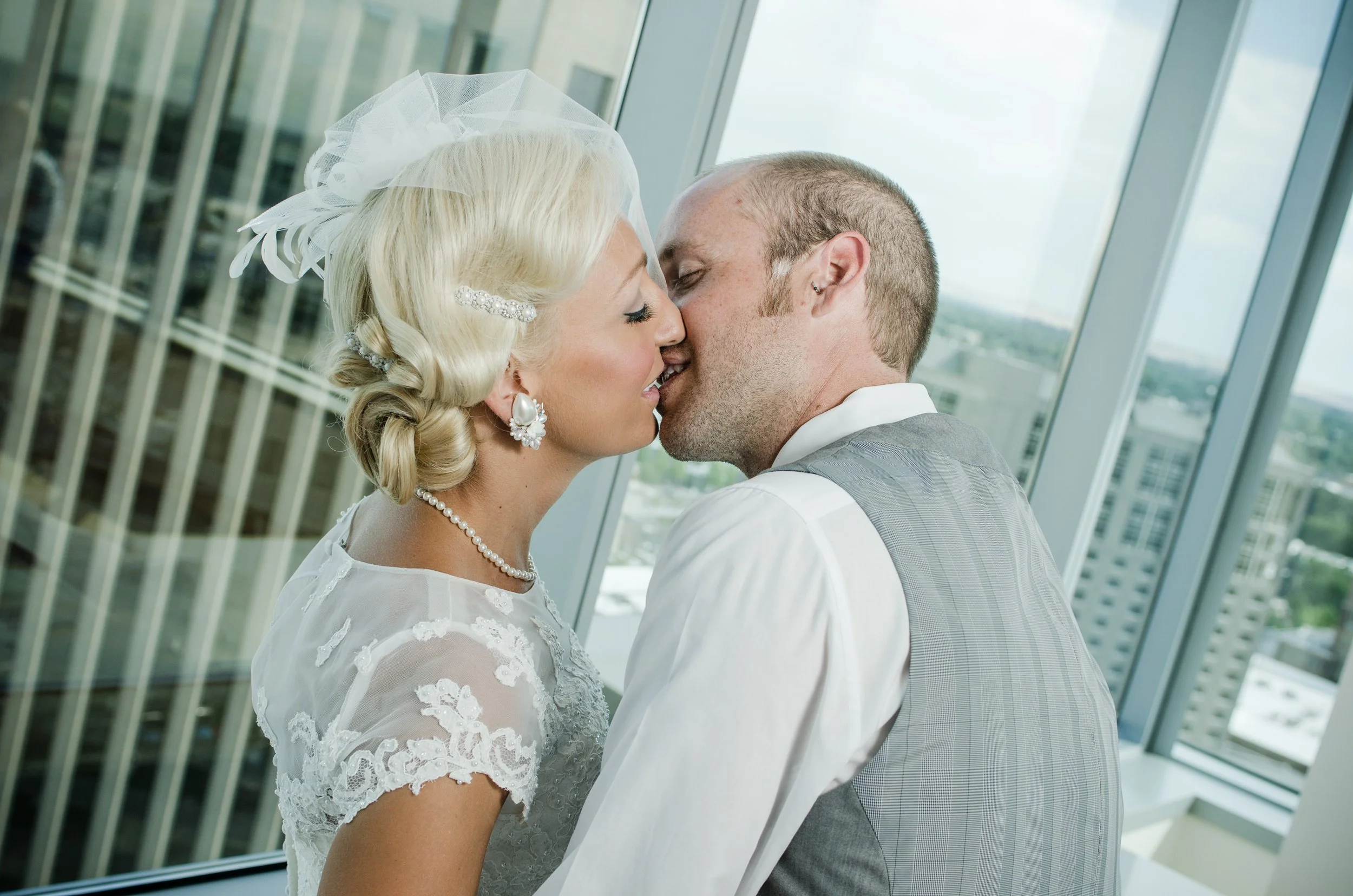 A bride and groom share a kiss indoors with large windows and a cityscape view in the background.