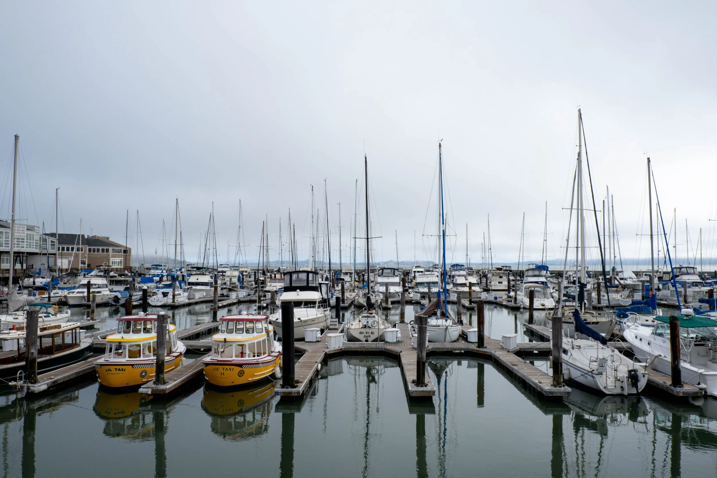 A marina with numerous sailboats and motorboats docked, some with colorful covers, under a cloudy sky. Buildings line the waterfront, and the water reflects the boats and structures.