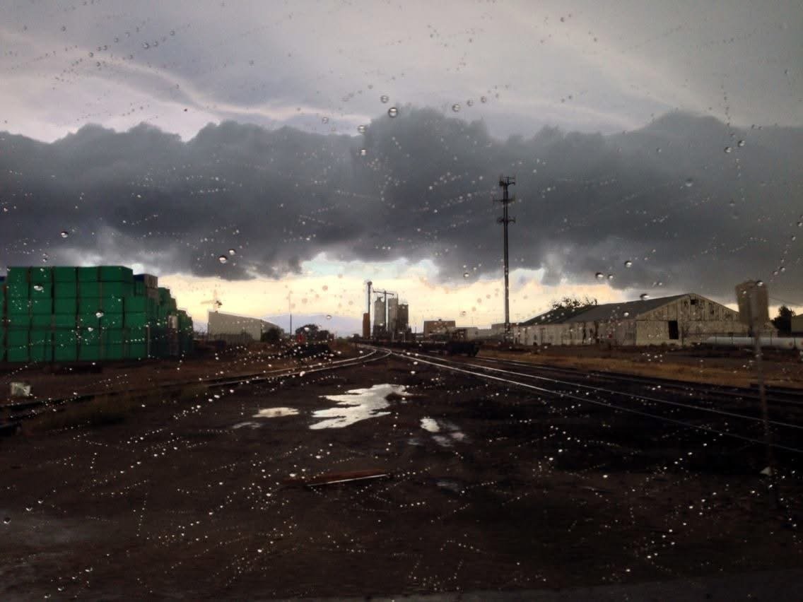 Dark, cloudy sky with rain droplets on a window, with train tracks, water puddles, and industrial buildings in the background.